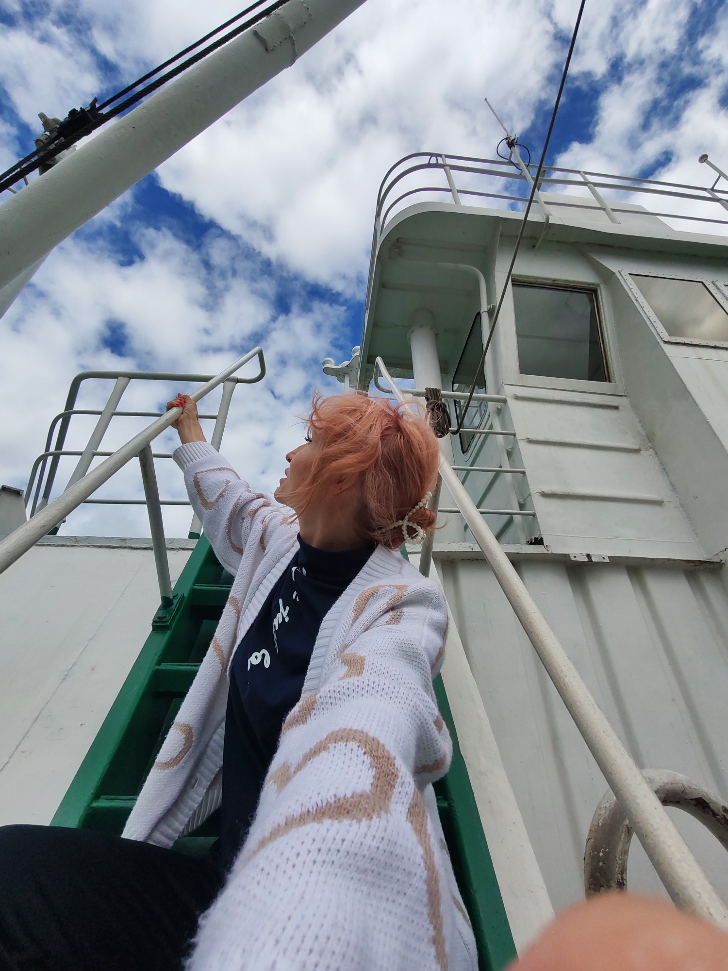 A low-angle, wide shot captures a person with bright orange hair reaching up to a white railing on the deck of a white vessel. The person is wearing a white cardigan with a tan geometric pattern over a dark shirt and black pants. Their right hand grips the railing, and they are looking upwards. To the left, green stairs lead upwards. The vessel's structure, including a white railing and part of a cabin with windows, is visible. Above, a cloudy blue sky is seen. The scene is set on the deck of a ship, likely in or near the city of Vytegra, Russia, given its proximity to the Vytegra River and Lake Onega, common waterways for vessels.