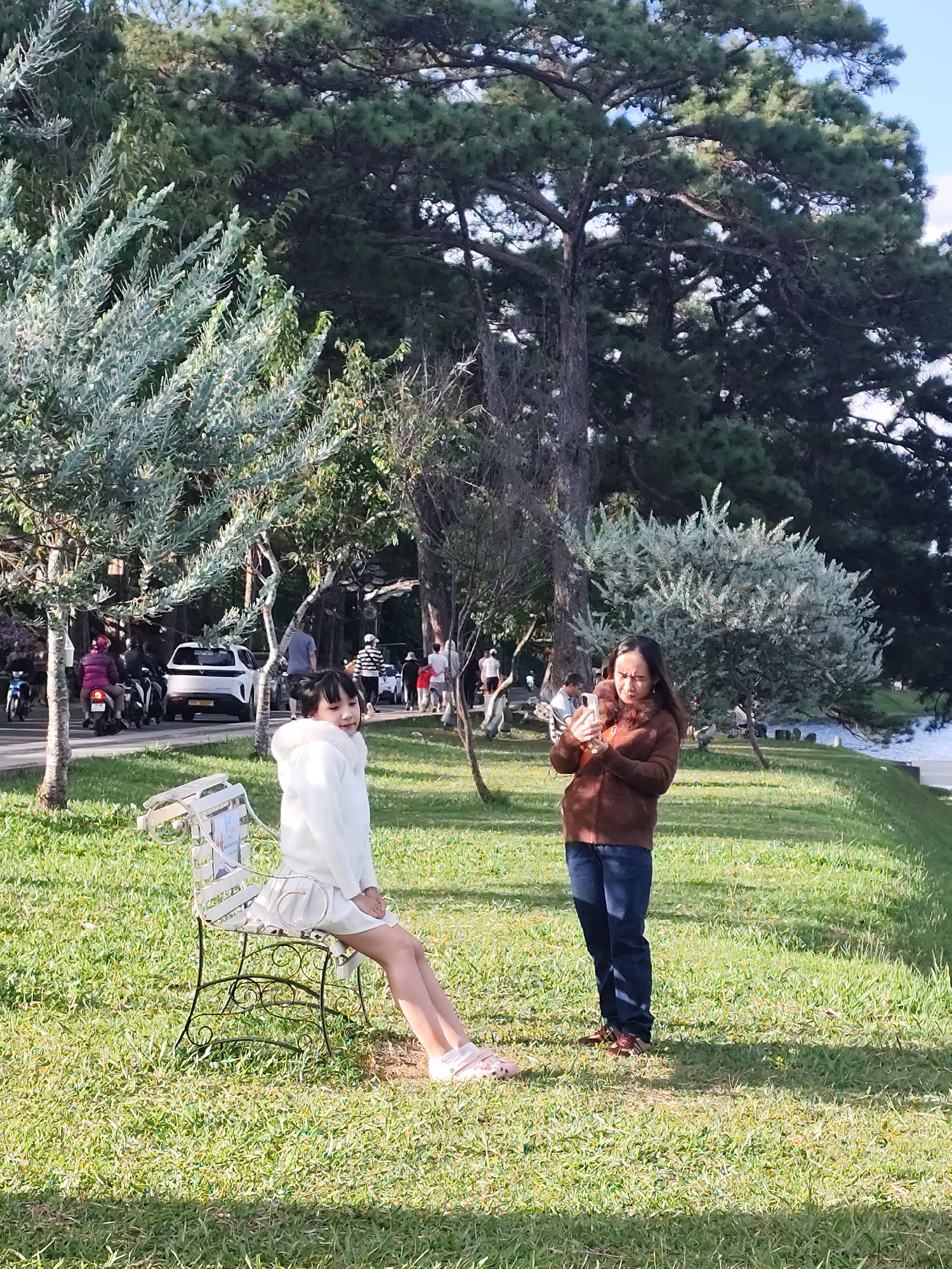 The image depicts a sunny outdoor scene in Xuân Hương Lake park, Đà Lạt, Vietnam. In the foreground, a young girl with dark hair, wearing a white hooded jacket and skirt, is seated on a decorative metal bench. She is looking to her right, with a slight smile. Next to her, a woman in a brown jacket and blue jeans is standing, holding a mobile phone in her hands and looking down at it. Behind them, a paved road is visible with traffic, including motorcycles and a white SUV. The background features lush green grass, various trees including pine trees, and a body of water, likely Xuân Hương Lake, with people in the distance. The overall atmosphere is one of a casual outdoor gathering in a park setting.