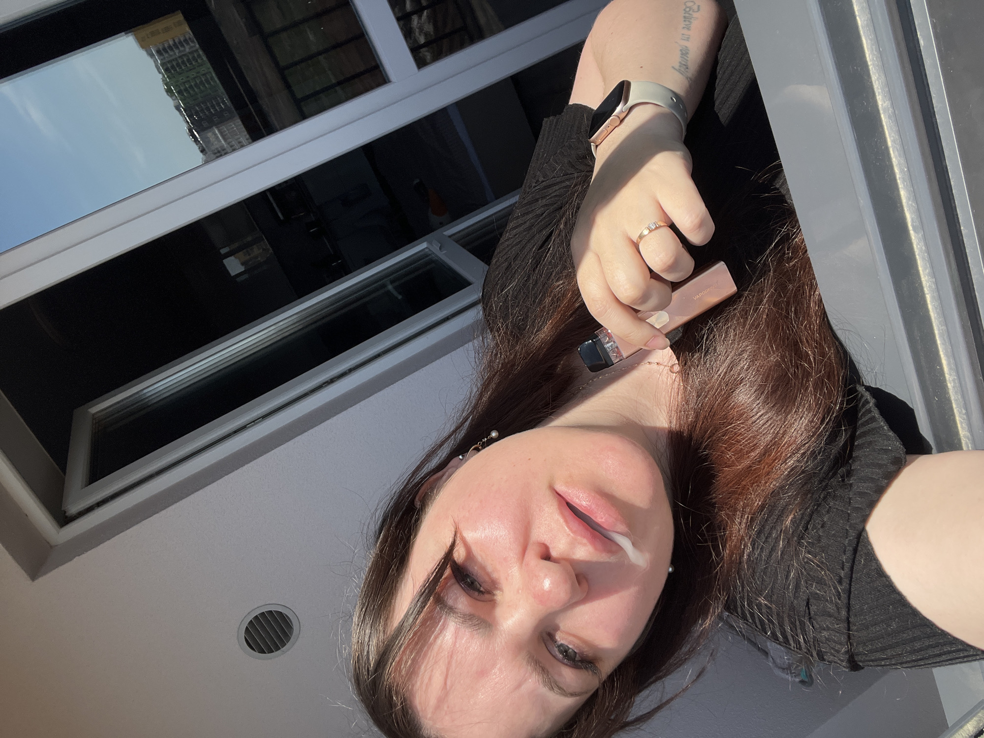 A selfie from an individual with long brown hair, pale skin, and light-colored eyes. The individual is holding a Vaporesso electronic cigarette. A white liquid appears to be emanating from the corner of her mouth. She is wearing a ring on her left hand and a smart watch. The background features a window with a reflection of a building. Location: Unknown.