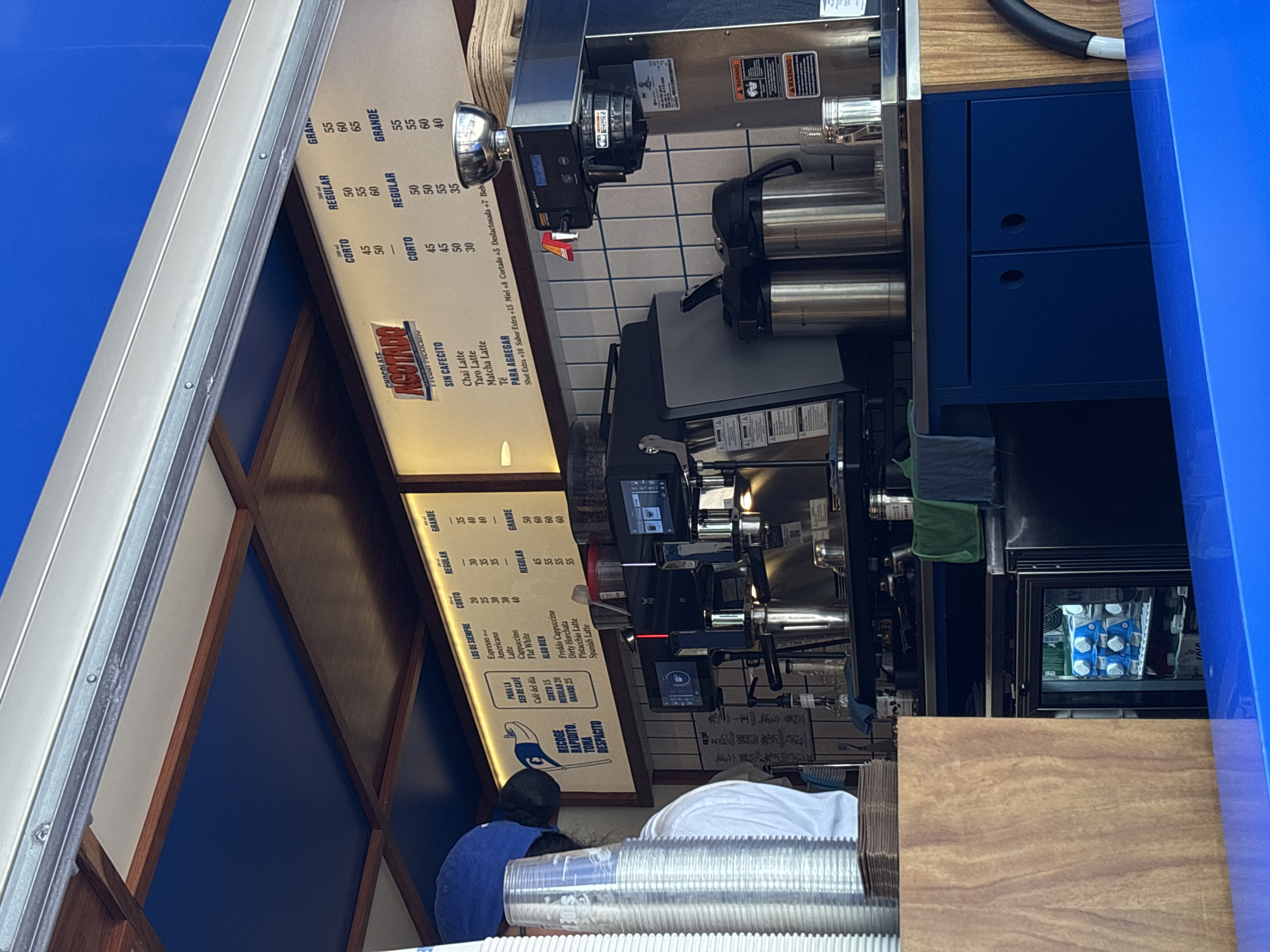 A coffee stand in Mexico City, Mexico. The stand has a menu board listing coffee and tea drinks with prices in pesos. Two people are near the counter, and the stand is equipped with coffee-making machinery, a refrigerator, and disposable cups. The stand is primarily blue, and the overall environment appears to be an outdoor or semi-outdoor setting.