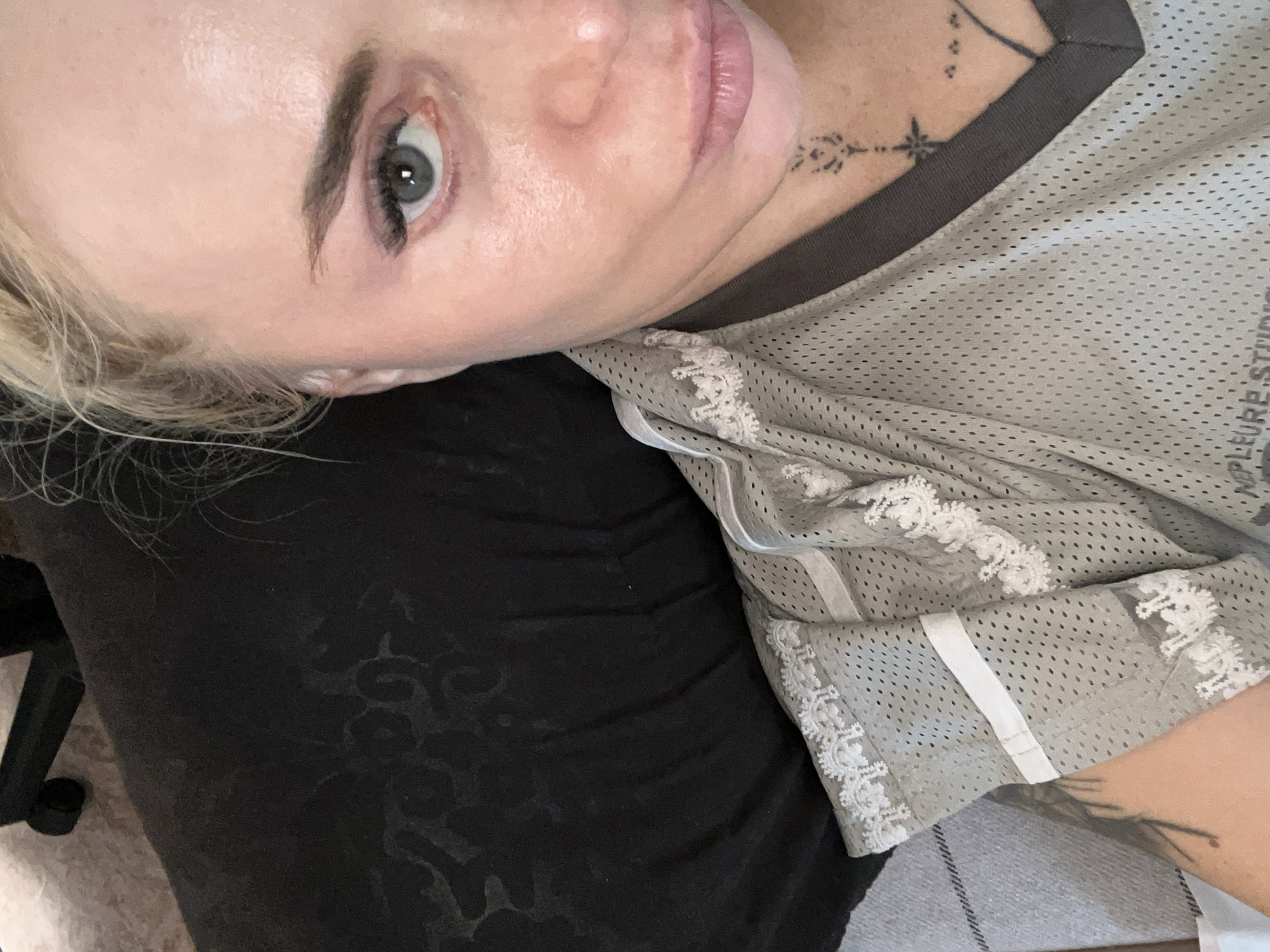 A close-up, high-angle medium shot shows the right side of a person's face and upper body, with the camera pointed downwards. The person has fair skin, blonde hair pulled back loosely, and is wearing light grey mesh athletic wear with white lace trim. They have makeup on, including eyeliner and mascara, and a tattoo on their neck. The background is dark and out of focus, with hints of a black fabric pattern and a grey striped surface. The lighting appears to be from above, creating highlights on the person's face. The scene is indoors, with no specific city or location details visible.