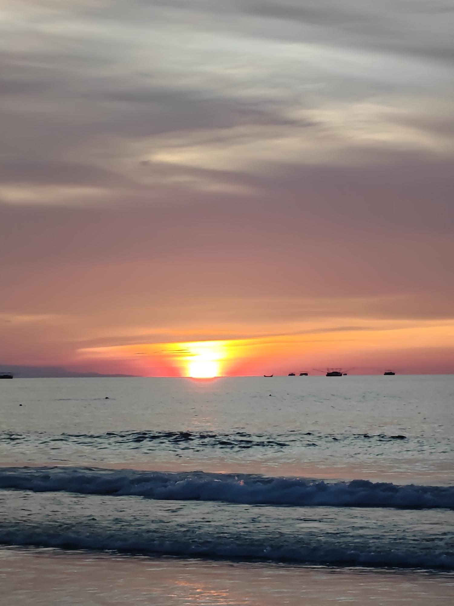 This image captures a serene sunset over the ocean at Curehtunong, Indonesia. The sky is a canvas of soft, diffused colors, transitioning from pale grey and dusty rose at the top to vibrant hues of orange and red as it meets the horizon. Wispy clouds add texture to the sky, catching the last rays of the sun.
The sun, a brilliant orb of yellow and red, is positioned just above the horizon, casting a warm glow across the water. The ocean itself is calm, with gentle waves rolling towards the shore. The foreground features a close-up view of these waves, their white foam contrasting with the darker, deeper blues and grays of the water. The wet sand at the very bottom of the frame reflects the sunset's colors, adding a subtle pinkish hue.
In the distance, several small boats are visible on the water, silhouetted against the vibrant sky. These boats suggest human activity and perhaps local fishing or transportation. The overall atmosphere is one of tranquility and natural beauty, evoking a sense of peace and the end of a day. The time of day is clearly sunset, indicated by the sun's position and the warm, rich colors in the sky and on the water. There is no visible text in the image.