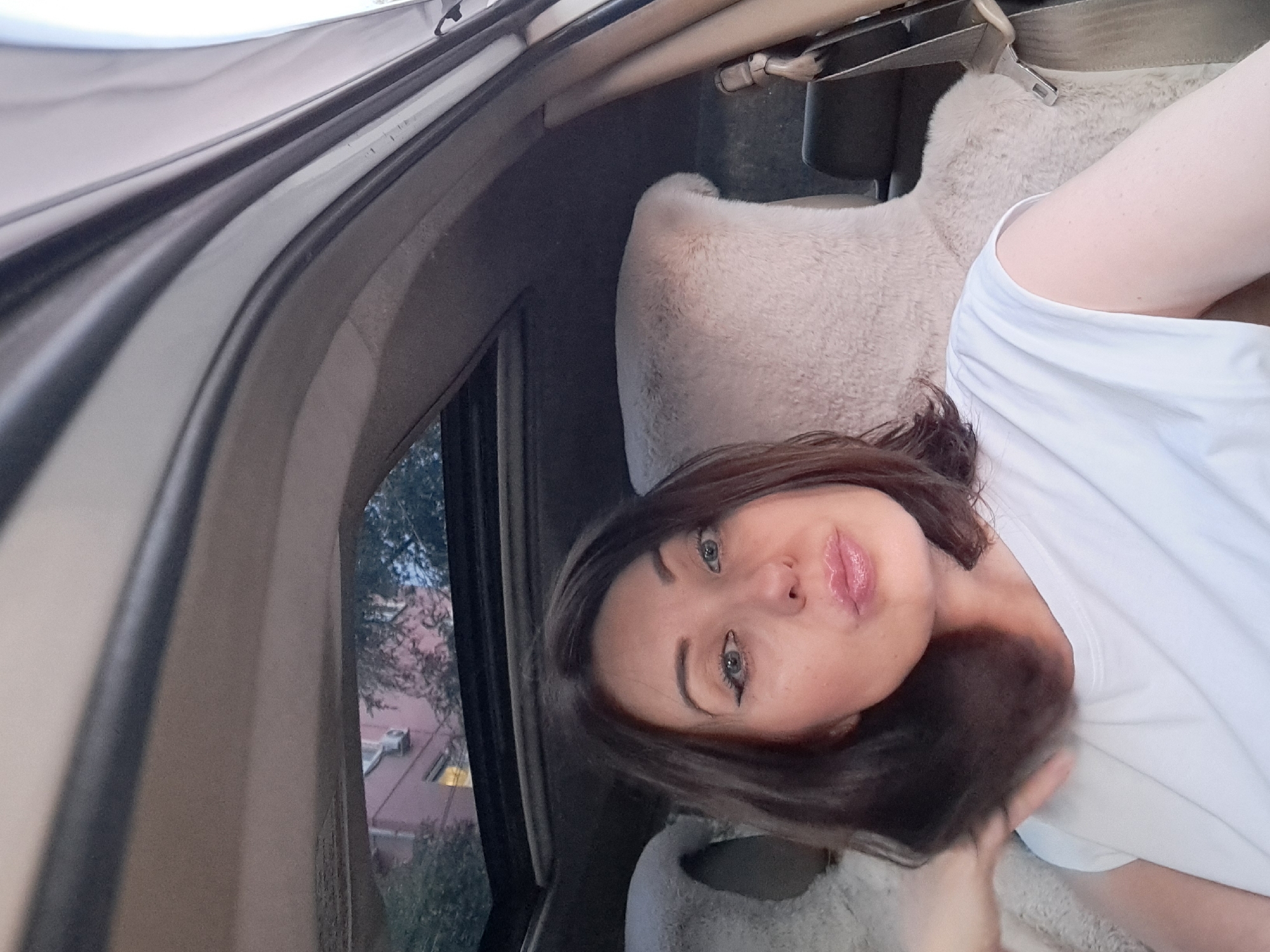 A woman with dark brown hair and light blue eyes is in the passenger seat of a car, looking at the camera and making a kiss face. She is wearing a white t-shirt. The car's sunroof is open, revealing a building with a pink facade and trees outside. The seats of the car are covered in a light-colored, fluffy material. The image was taken in Irkutsk, Russia.