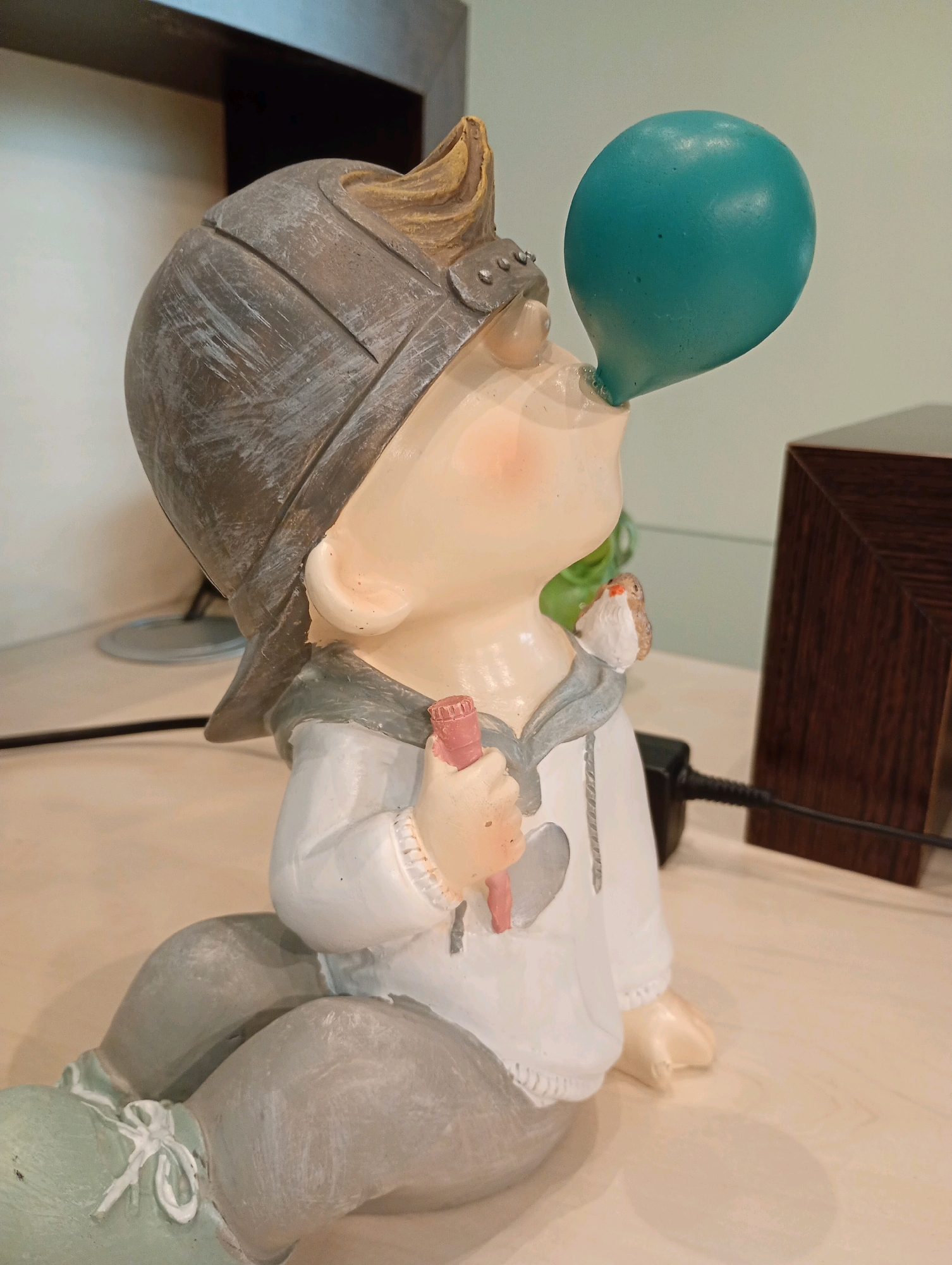 A close-up, eye-level, side-view shot captures a ceramic figurine of a young child wearing a helmet and a white hoodie, sitting on a light-colored surface. The figurine is blowing a turquoise balloon, which protrudes from its mouth. A small, brown and white bird is perched on the figurine's shoulder. The child's right hand is holding a pink, cylindrical object. The background is blurred, showing a dark wooden object to the right, a white wall, and what appears to be part of a shelf or furniture. The scene is indoors, likely on a desk or shelf. There is no discernible connection to the city of Bad Sachsa, Germany.