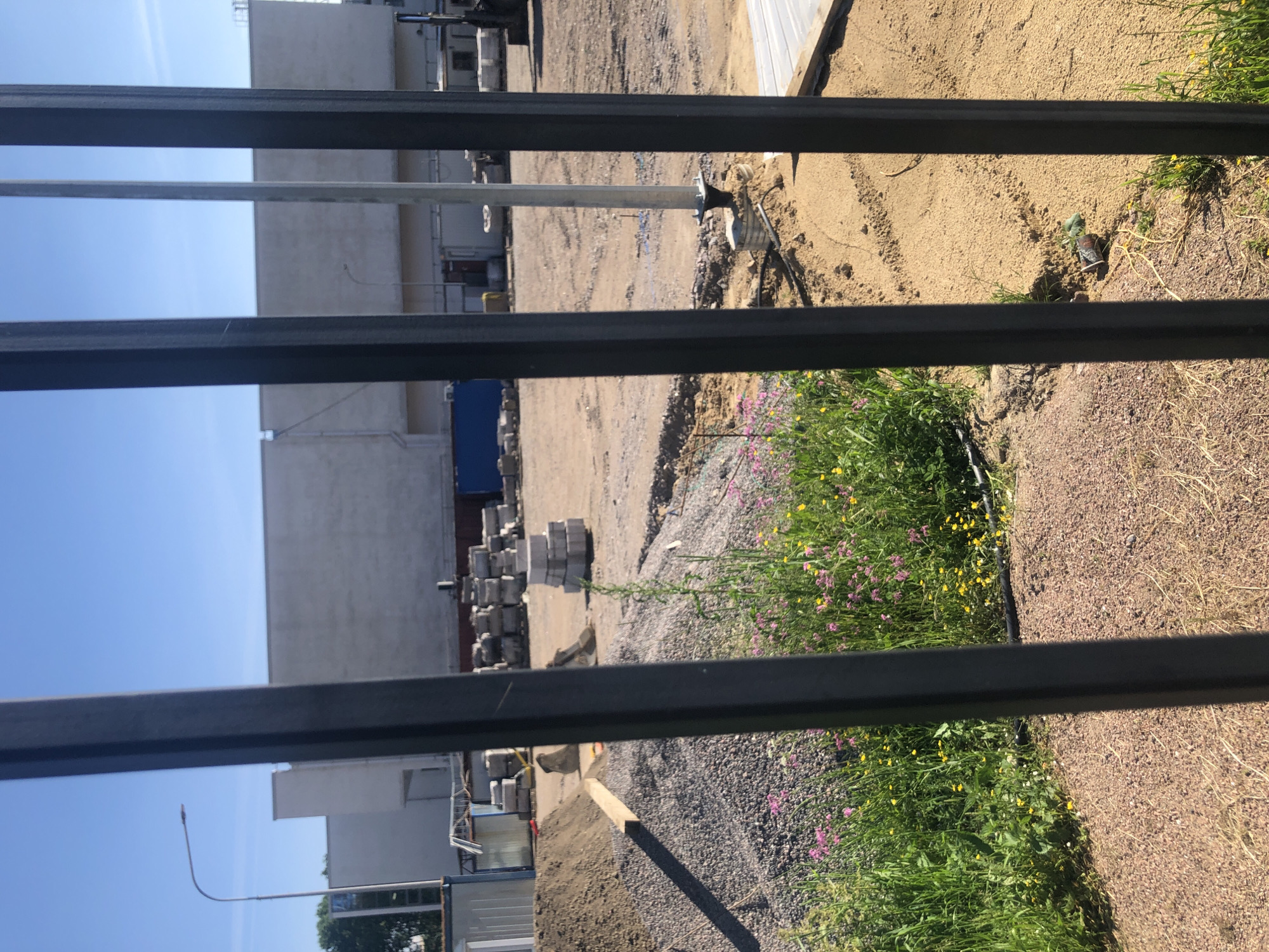 This image shows a construction site behind a black metal fence. In the background, there is a large, light-colored building. In the middle ground, there are piles of gravel and sand, stacks of concrete blocks, and some construction equipment. In the foreground, there are wildflowers and grass growing. The sky is blue and clear. The location is the construction site of the "Lenta" hypermarket on Kolomyazhsky Prospekt in Saint Petersburg, Russia.