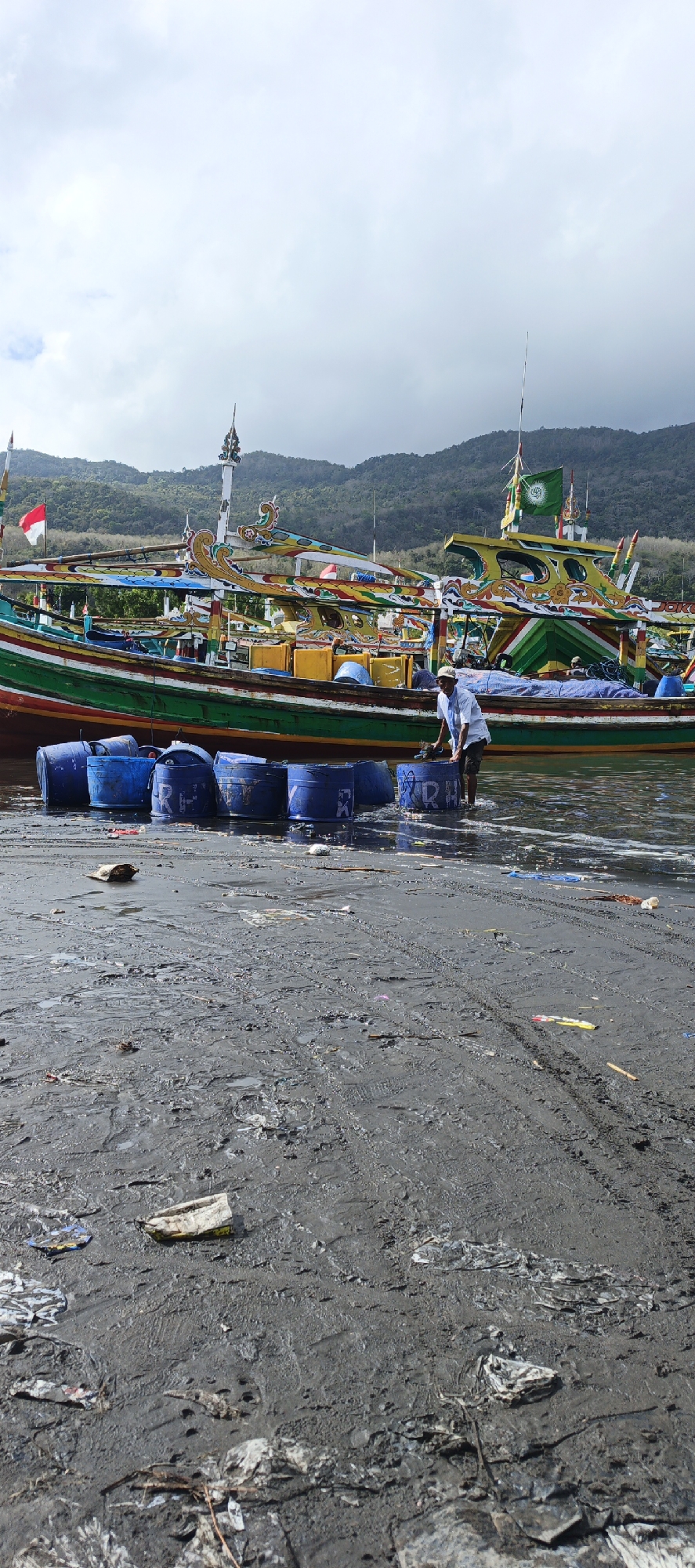 In the city of Puger, Indonesia, a man is wading in shallow water near a line of blue plastic barrels. He is wearing a white long-sleeved shirt, dark pants, and a light-colored cap. The barrels are positioned in the water in front of several vibrantly painted fishing boats. The boats are adorned with colorful decorations, flags, and intricate carvings. Behind the boats, a lush green hillside rises under a cloudy sky. The foreground of the image shows a muddy, debris-strewn shoreline.