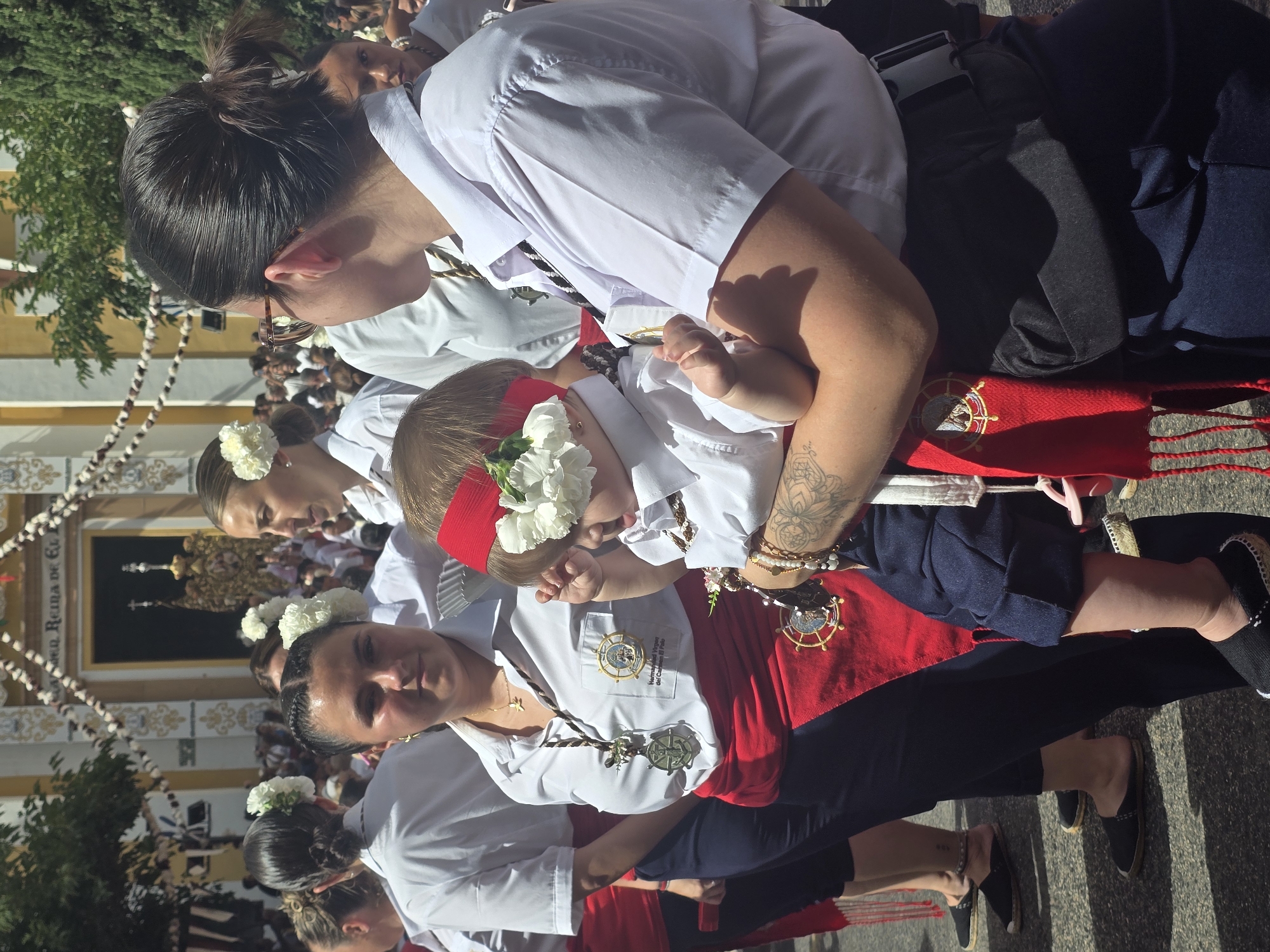 This media file depicts a group of people, predominantly women and a baby, participating in a religious procession likely in Málaga, Spain. The subjects are dressed in white shirts and many are adorned with white flowers in their hair. Some are wearing red sashes with embroidered emblems, and several individuals have necklaces with medals. The baby, at the center of the image, is also wearing a white shirt and a red band with white flowers around its head.
The scene appears to be outdoors on a sunny day, given the strong shadows and bright lighting. The background suggests an urban setting, possibly a street lined with buildings, one of which has a grand facade with ornate decorations. There is also a glimpse of greenery, likely trees. The people are walking in a procession, indicated by their organized formation and the presence of what appears to be a religious float or display in the background, partially obscured. The atmosphere seems solemn yet celebratory, typical of a religious festival.
The overall impression is one of community and shared tradition, with a focus on religious devotion and cultural heritage. The expressions on the faces of the adults suggest a mix of focus and calm, with the baby representing innocence and future generations within the tradition.
Visible text includes "Reina de Co..." which likely refers to "Reina de la Costaleros" or a similar religious title associated with a queen or patron saint, and possibly "Hermandad" which means brotherhood or fraternity, common in Spanish religious confraternities. There is also text on a badge worn by one of the individuals which reads "Hermandad Virgen del..." followed by more obscured text.