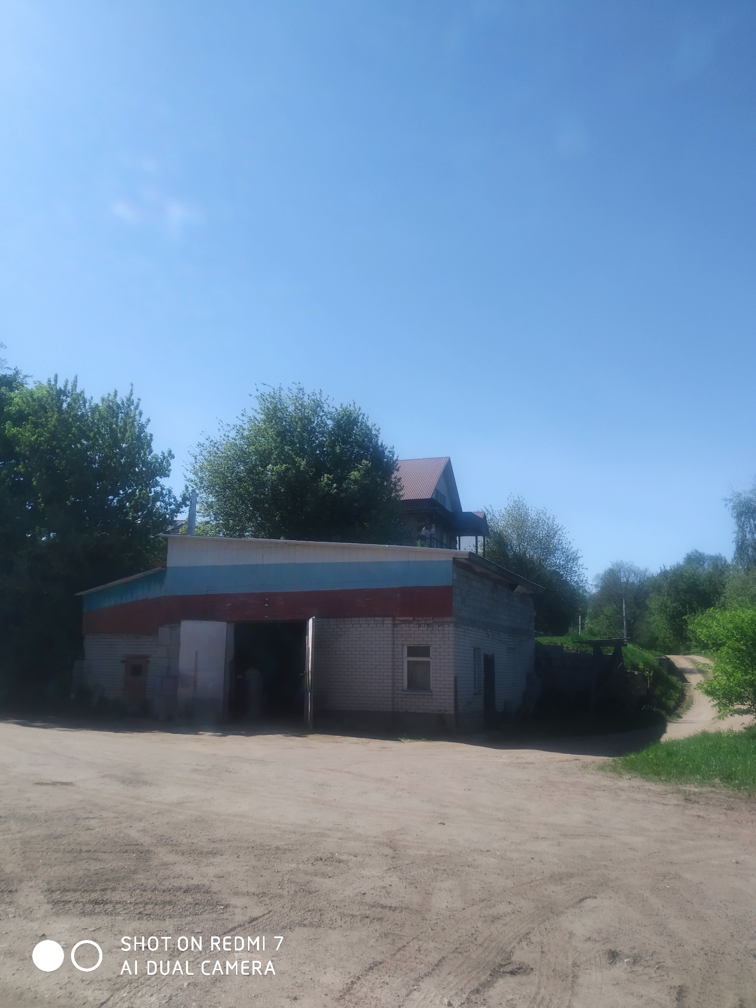 The image depicts a single-story, brick building in Ochansk, Russia. The building has a large open doorway and a smaller window. The roof is painted with the colors of the Russian flag. A dirt road leads past the building, with trees and greenery in the background. In the distance, a two-story house can be seen. The sky is blue. The ground is dirt and appears to be dry. There are no people visible in the image.