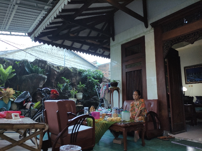The scene is a partially covered patio of a home in Mojokerto, Indonesia (latitude: -7.459, longitude: 112.447). It is late afternoon, and the sky is clear. A woman sits on a couch facing the camera, dressed in a printed brown and white blouse, and black pants. She appears to be resting and looking at the camera. Her left foot is resting on the floor and her right foot is on a step stool. There is a small table in front of her with a bowl of food. Behind her is an intricately carved wooden door leading to a doorway with a traditional Indonesian painting hanging on the wall. To the left of the woman is a wooden chair, and behind the chair is a bicycle. The patio is enclosed by a white wall with a trellis-like top. The roof is made of wood, and there are several plants and flowers growing in pots in the background. The patio is decorated with traditional Indonesian furnishings. The scene is peaceful and relaxed.