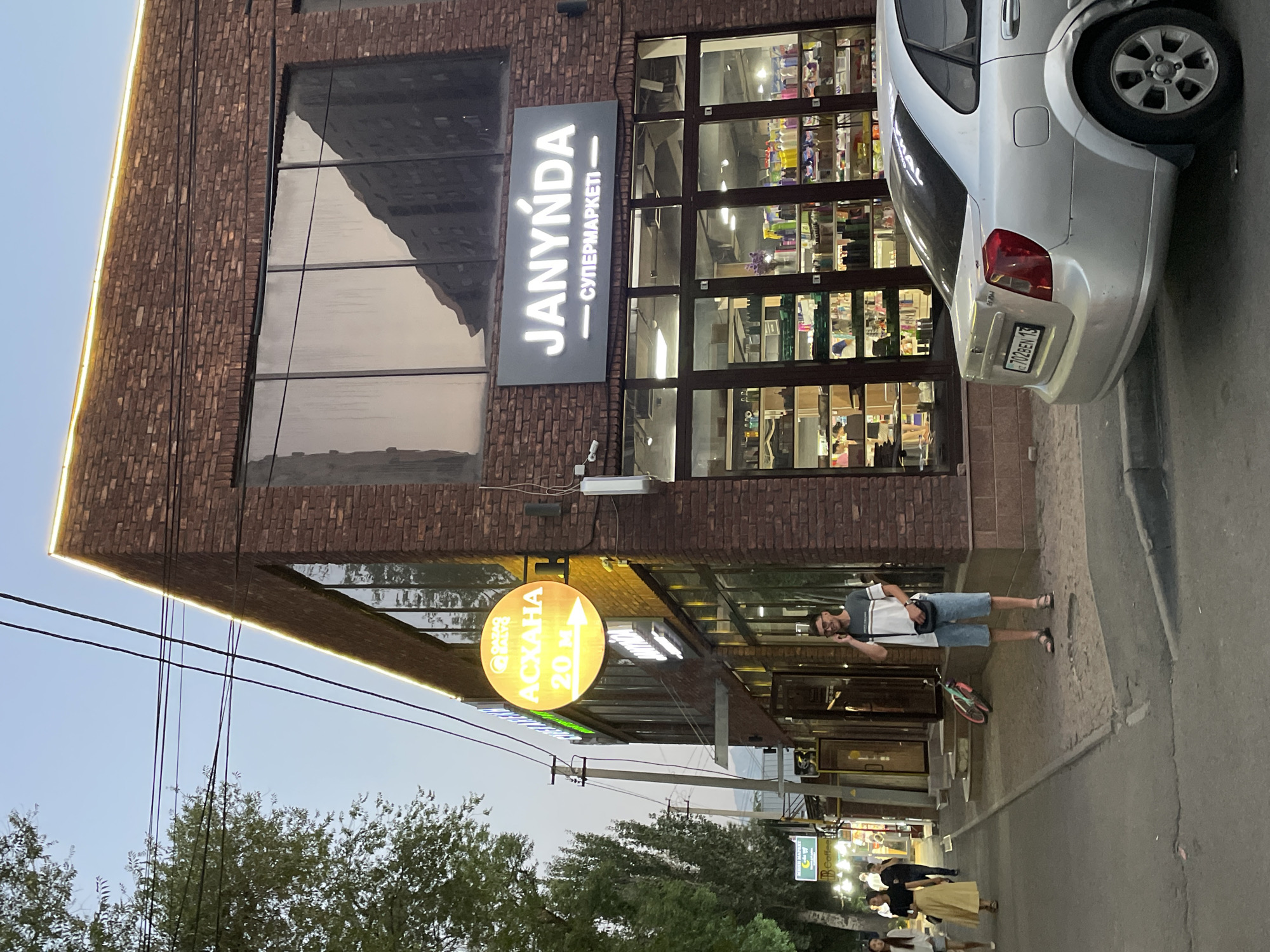 The image depicts a street scene in Almaty, Kazakhstan, likely during the late afternoon or early evening. A modern brick building, featuring large glass windows and a prominent illuminated sign reading "JANYNDA - СУПЕРМАРКЕТ", dominates the foreground. The sign is in a sans-serif font, with "JANYNDA" in large, white capital letters and the Russian text "СУПЕРМАРКЕТ" below it.
To the right of the building, a silver sedan is parked, its rear visible. The car has a license plate with the inscription "702BEN 13". A person wearing a t-shirt, shorts, and sandals is standing on the sidewalk in front of the building, looking towards the camera and gesturing with their hand. A bicycle with a pink frame is leaning on a wall nearby.
Further down the street, another illuminated circular sign, predominantly orange with white text, hangs from a pole. This sign appears to be an arrow pointing to the right, with the text "АСКАHA 20 M" (likely indicating "Ascena 20 meters" in Russian). The entrance to the supermarket is visible, with people walking in and out.
In the background, more people are seen walking along the sidewalk, and glimpses of other shop fronts suggest a commercial area. Trees line the street, and power lines crisscross the sky, which is a pale blue, indicating dusk or a clear day. The overall atmosphere seems casual and urban.