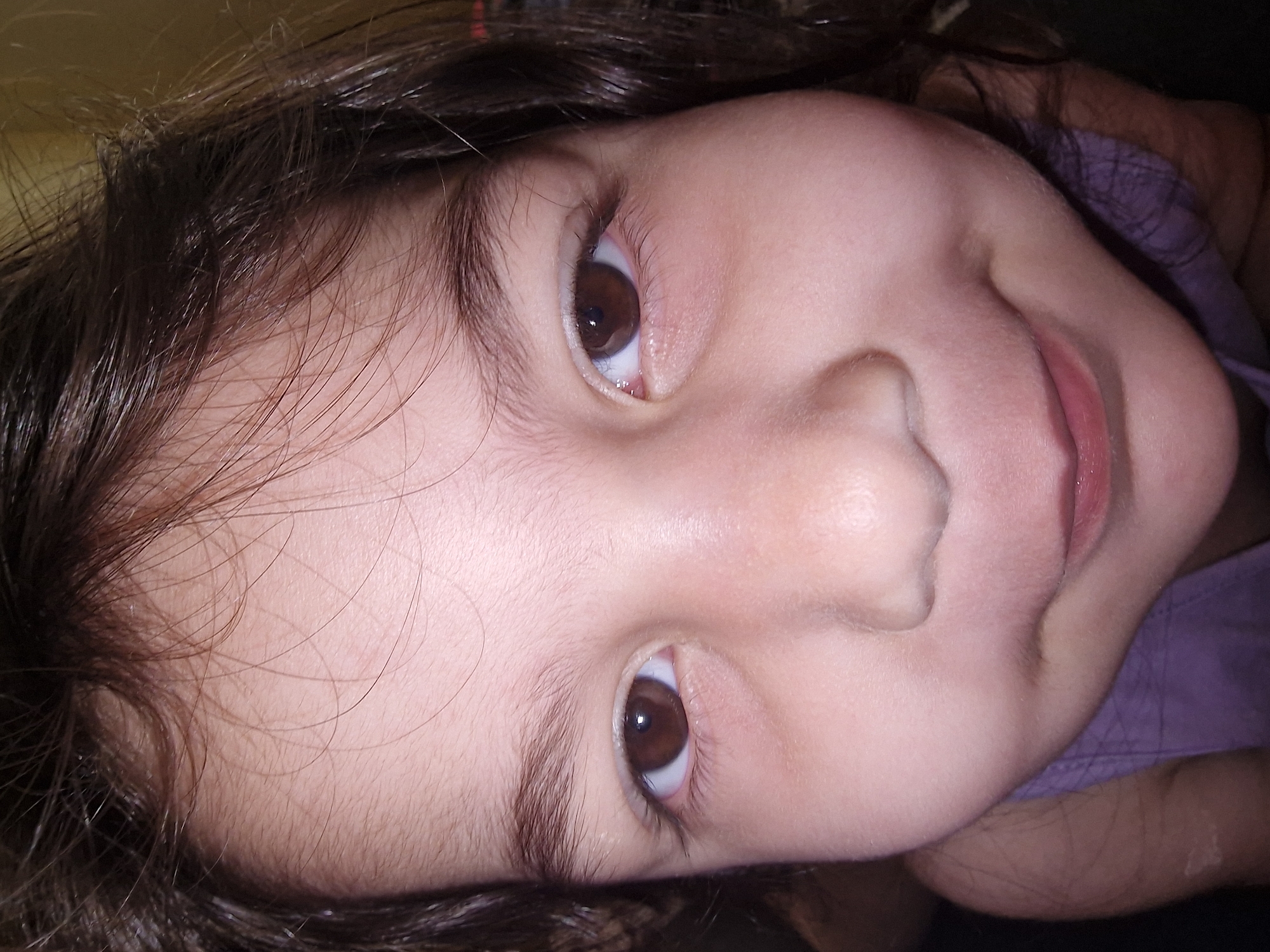 A close-up, angled shot of a young child's face, featuring brown hair and brown eyes. The child's skin is fair, and they are wearing a purple garment. The location is not specified.