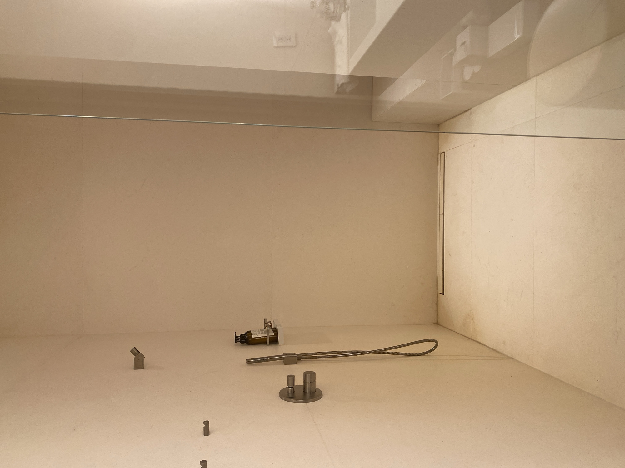 This image appears to be a photograph taken inside a minimalist, possibly institutional or museum-like space. The scene is dominated by light-colored walls with subtle vertical seams, suggesting large panels or tiles. A horizontal metal rod spans the upper portion of the frame, intersecting where the side walls meet the back. A glass ceiling or barrier is visible in the upper portion of the image.
On the floor, several small, metal objects are arranged. From left to right: a small angled piece, a collection of what appears to be an amber colored liquid dispensing bottle next to a complex piece of machinery, then a flexible metal tube attached to a cylindrical component, and two small cylindrical pieces protruding from a circular base. The walls are off-white with subtle seams, suggesting a simple background.
There are no people visible in the frame. There are no actions or interactions to describe based on the objects present.
There is no indication of a location in the city of El Caracol, Mexico, unless these objects are known to be an art installation or artifact exhibited in that specific locale.