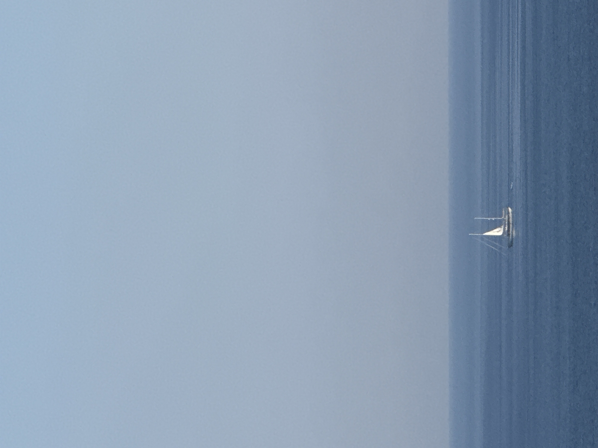 A solitary sailboat with white sails is depicted on a calm, deep blue sea. The horizon line is distinct, separating the water from a pale blue sky that gradually fades to a lighter shade at the top of the image. The sailboat is positioned in the lower center of the frame, appearing small against the vast expanse of water. There are no other vessels or landmasses visible. The scene is peaceful and serene, suggesting an open-water environment. The image does not provide any specific geographical markers or context related to the city of Boreti, Montenegro.