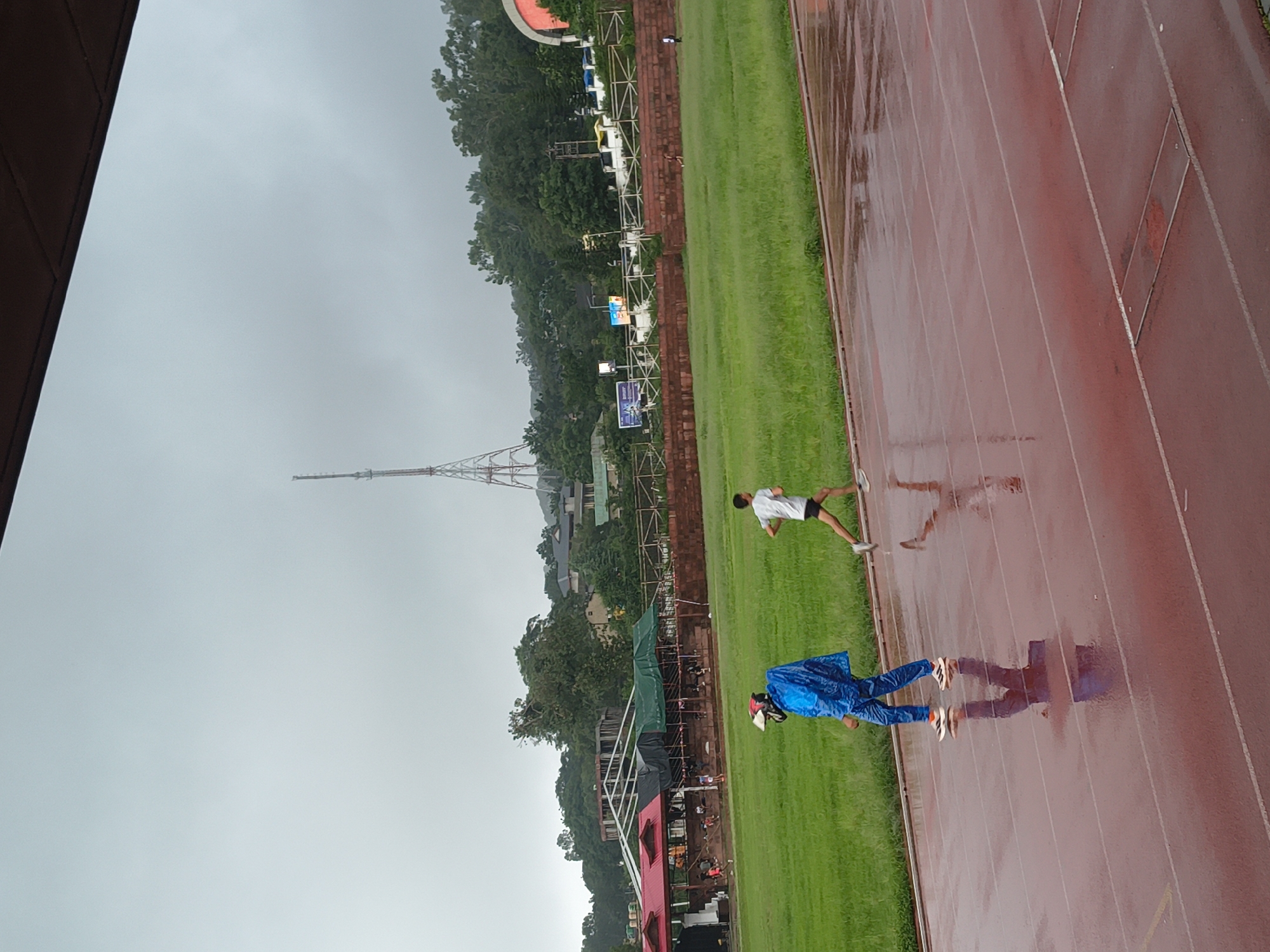 An overcast day at a stadium in Dharamshala, India. Two individuals are present on a wet, red running track. In the foreground, a person wearing a bright blue raincoat and pants walks towards the left, with their reflection visible on the slick track. To the right and slightly behind, a second person, a runner in a white t-shirt and black shorts, is captured mid-stride, also running towards the left. The background features a green field, stadium seating with people visible, trees, buildings, and a tall communication tower. The overall atmosphere suggests a rainy or recently rained-upon condition, impacting the stadium environment.