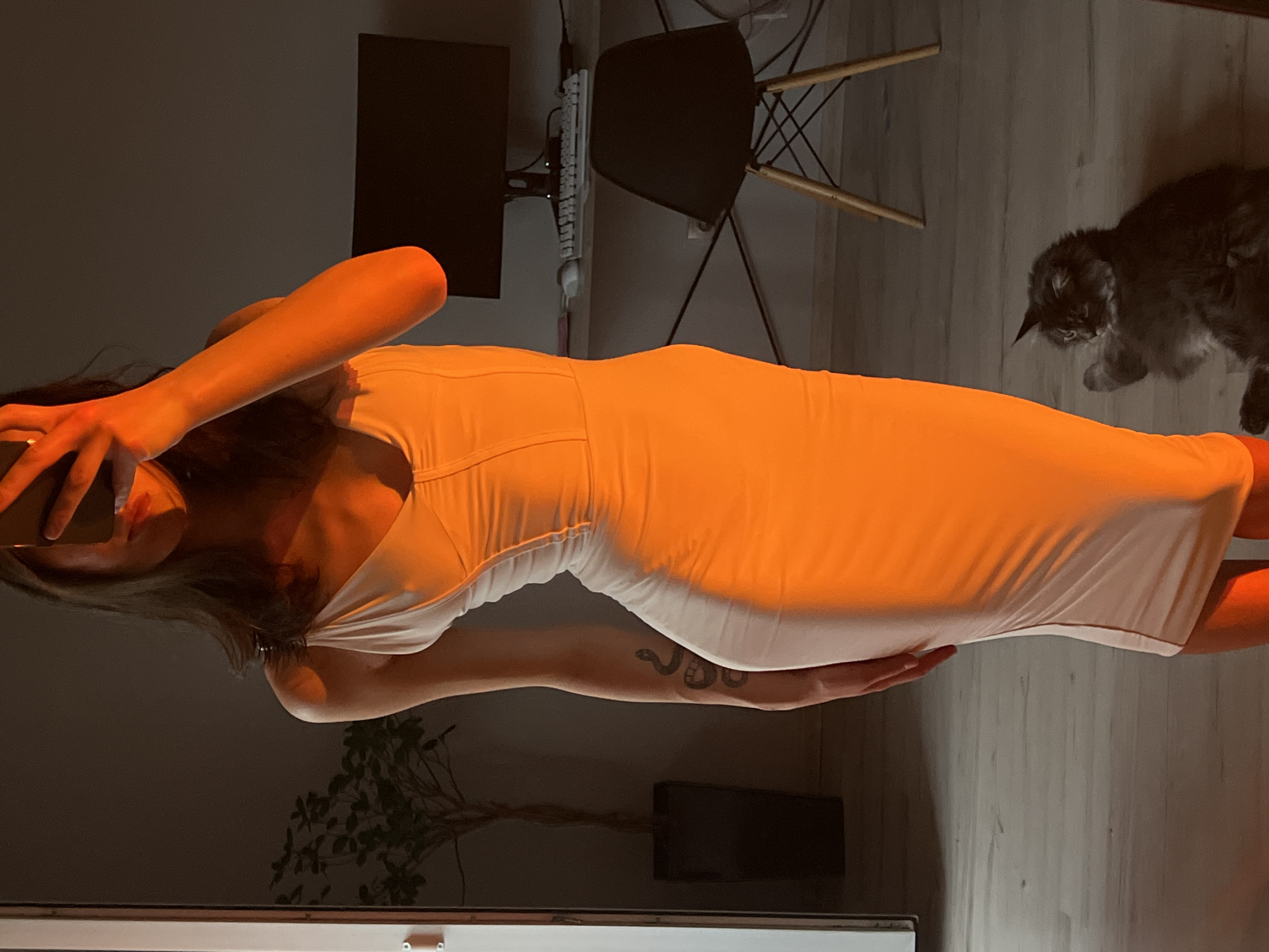 In a dimly lit room with wooden flooring, a woman in a white dress is captured in a close-up shot. Her left arm is raised, holding what appears to be a phone, suggesting she is taking a selfie or video call. The warm, orange glow from an unseen light source illuminates her skin and the fabric of her dress, creating a dramatic effect. A snake tattoo is visible on her forearm. In the background, a fluffy cat is perched on the floor, looking down, adding a touch of domesticity to the scene. A chair with a wooden frame and a black seat is positioned in the mid-ground, near a computer setup with a monitor and keyboard. The overall ambiance suggests a relaxed, intimate moment at home, possibly in the evening given the lighting.