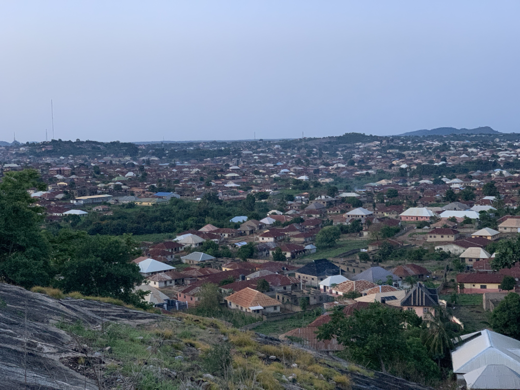 The image depicts a dense urban landscape in Kabba, Nigeria, viewed from an elevated position. The scene is dominated by closely packed buildings, predominantly single-story residential structures with diverse roof colors and materials including terracotta and metal sheeting. Interspersed among the houses are patches of green vegetation, with trees and shrubbery evident in both the foreground and scattered throughout the city. A rocky, grass-covered terrain forms the immediate foreground, suggesting the vantage point is from a hillside or outcrop. In the distance, the city extends towards the horizon, where a line of hills is visible. The sky is a pale blue, indicating either early morning or late afternoon light. Tall communications towers are visible in the distance. The overall impression is of a sprawling, densely populated urban environment set within a natural, hilly landscape.