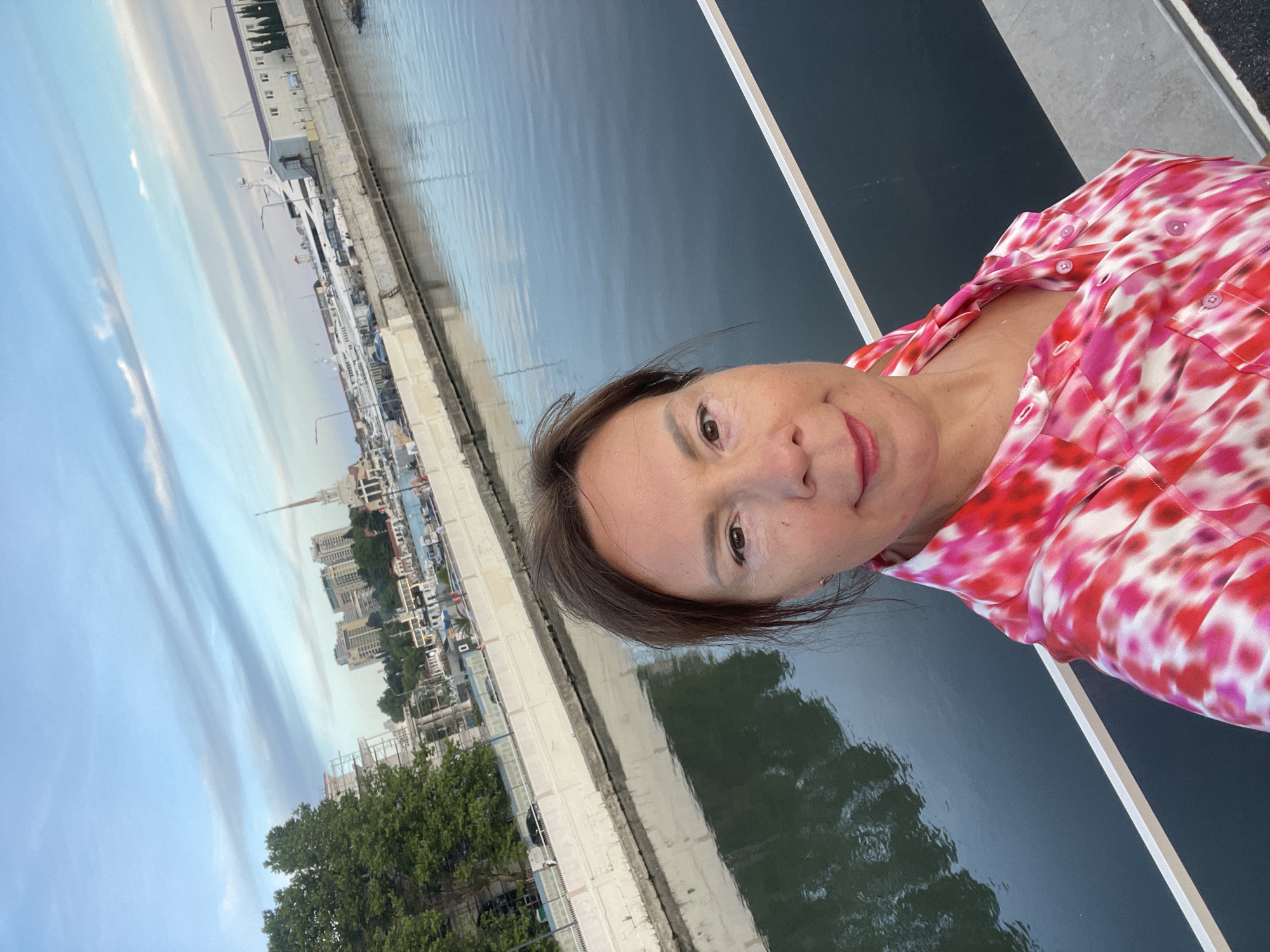 A woman in a pink and white patterned shirt takes a selfie on a bridge overlooking a canal in Sochi, Russia. In the background, there are buildings, trees, boats, and a clear sky with clouds.