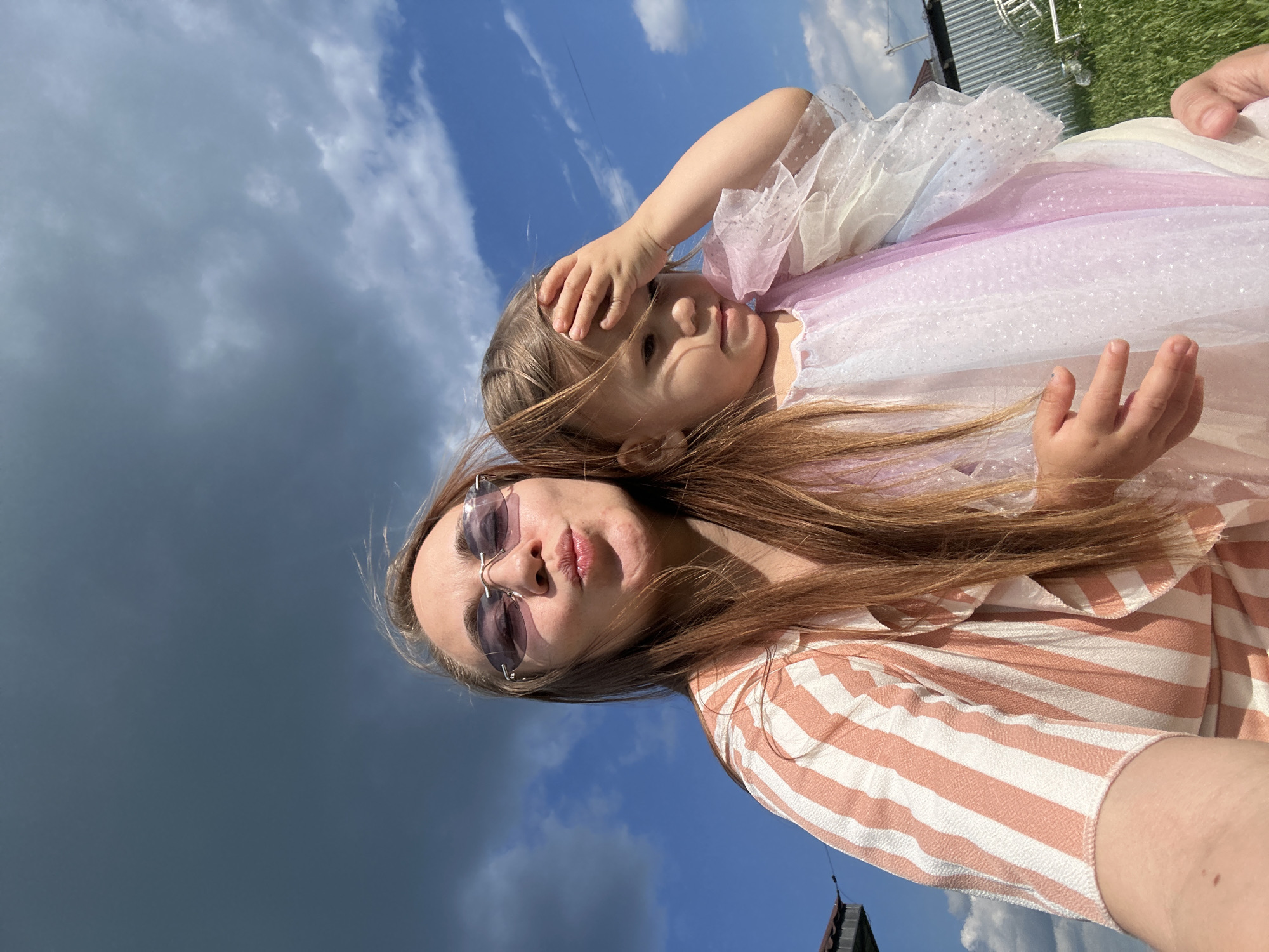 A woman wearing sunglasses and a peach and white striped shirt is taking a selfie outdoors. She is puckering her lips. A young girl in a pastel colored dress is standing next to her, with her hand on her forehead. The background is a blue sky with some clouds. The woman is in the foreground, and the girl is to her right. The scene appears to be in the city of Kharnavy, Russia.