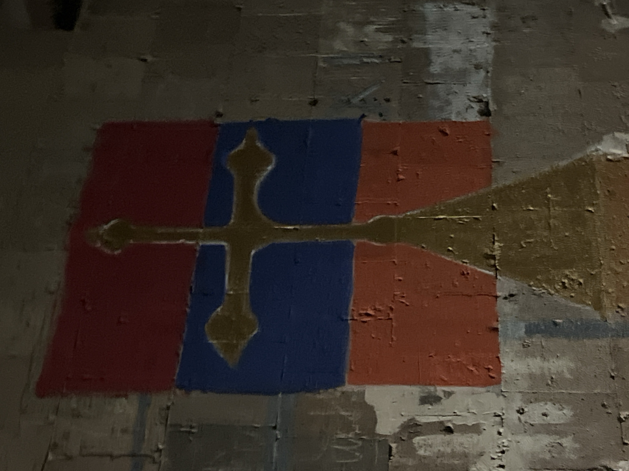 The image depicts what appears to be graffiti on a wall. The graffiti consists of a rectangular design divided into three vertical sections colored red, blue, and red. A gold-colored cross with decorative elements extends horizontally across the design. The cross has a flared base and top, and there is a golden spear like shape extending toward the right of the image. The background is a rough, uneven wall texture, suggesting it could be concrete or a similar material. The quality of the graffiti indicates it may be amateur or old.
I cannot determine the location in the city of Yerevan, Armenia, based solely on the visual information provided in the image.