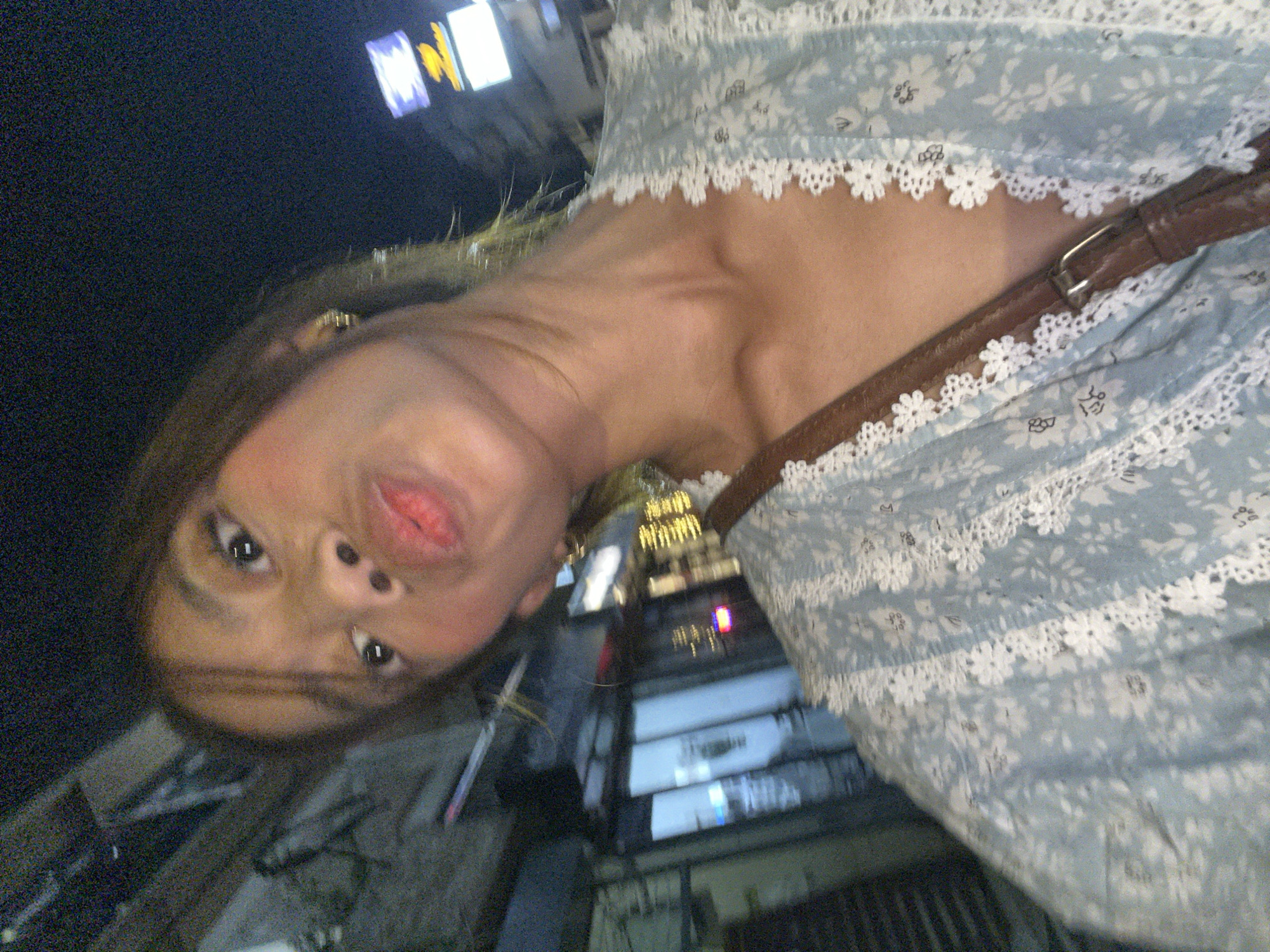 The image is a selfie taken at night, likely in a commercial area of Davao City, Philippines. The subject, a young woman, is posing with her lips puckered. She is wearing a light blue dress with a white floral pattern and white lace trim. A brown leather strap from a bag or purse is visible across her chest. The background is blurred and shows storefronts, some with illuminated signs, and what appear to be streetlights, suggesting an urban setting. The lighting is dim, consistent with nighttime.