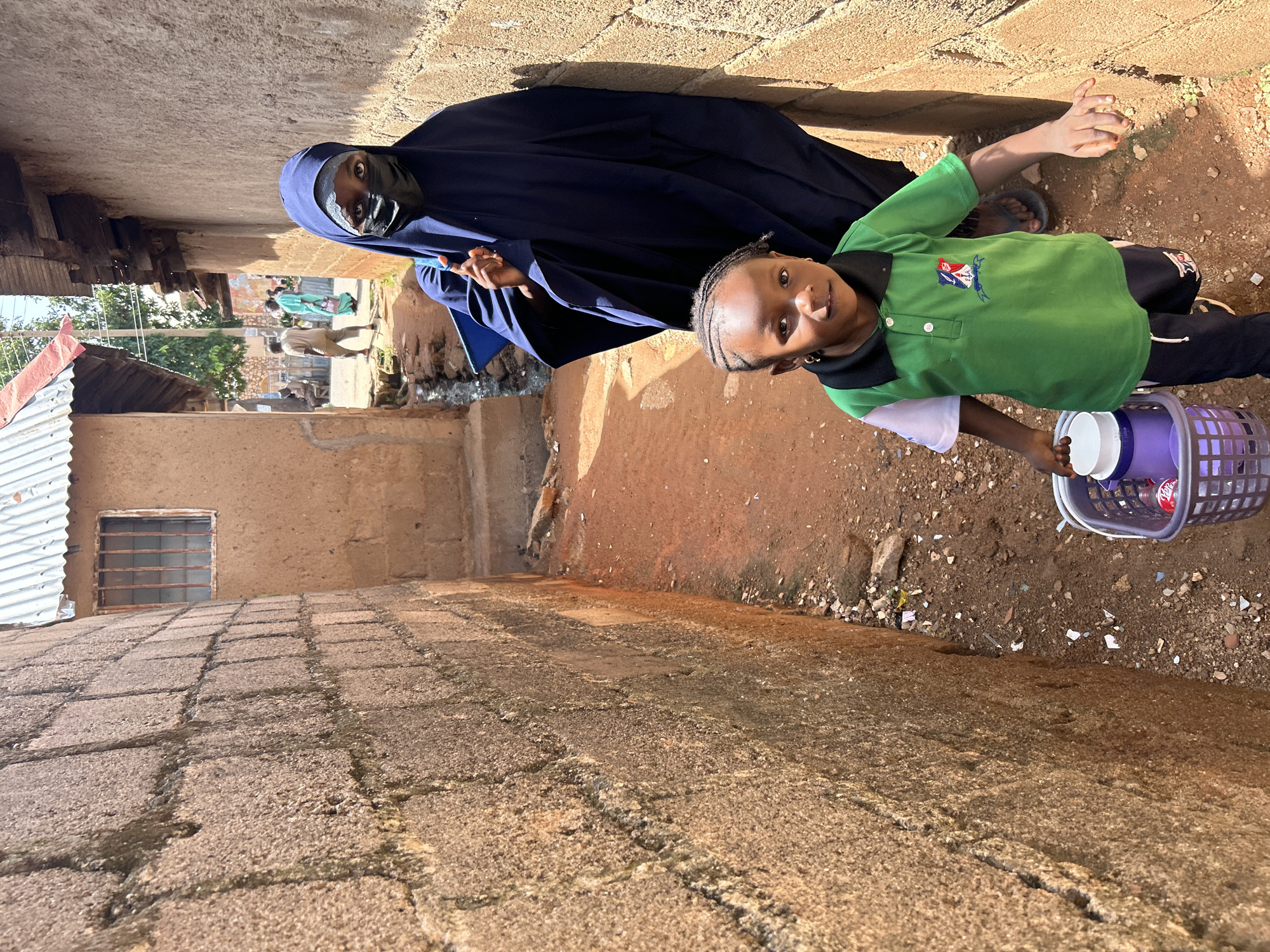 In a narrow alleyway in Jos, Nigeria, a young child in a green and white polo shirt and dark pants walks towards the camera, carrying a purple basket filled with items, including a white-capped container and a red-labeled bottle. Behind the child, a person wearing a dark blue hijab and a black face mask stands partially obscured by the child, their hand visible near the child's shoulder. The alley walls are constructed of rough, tan-colored bricks. In the background, other people are visible in the distance, as well as buildings with corrugated metal roofs and a small barred window. The ground of the alley is a mix of reddish-brown dirt and small stones, with some scattered debris.
