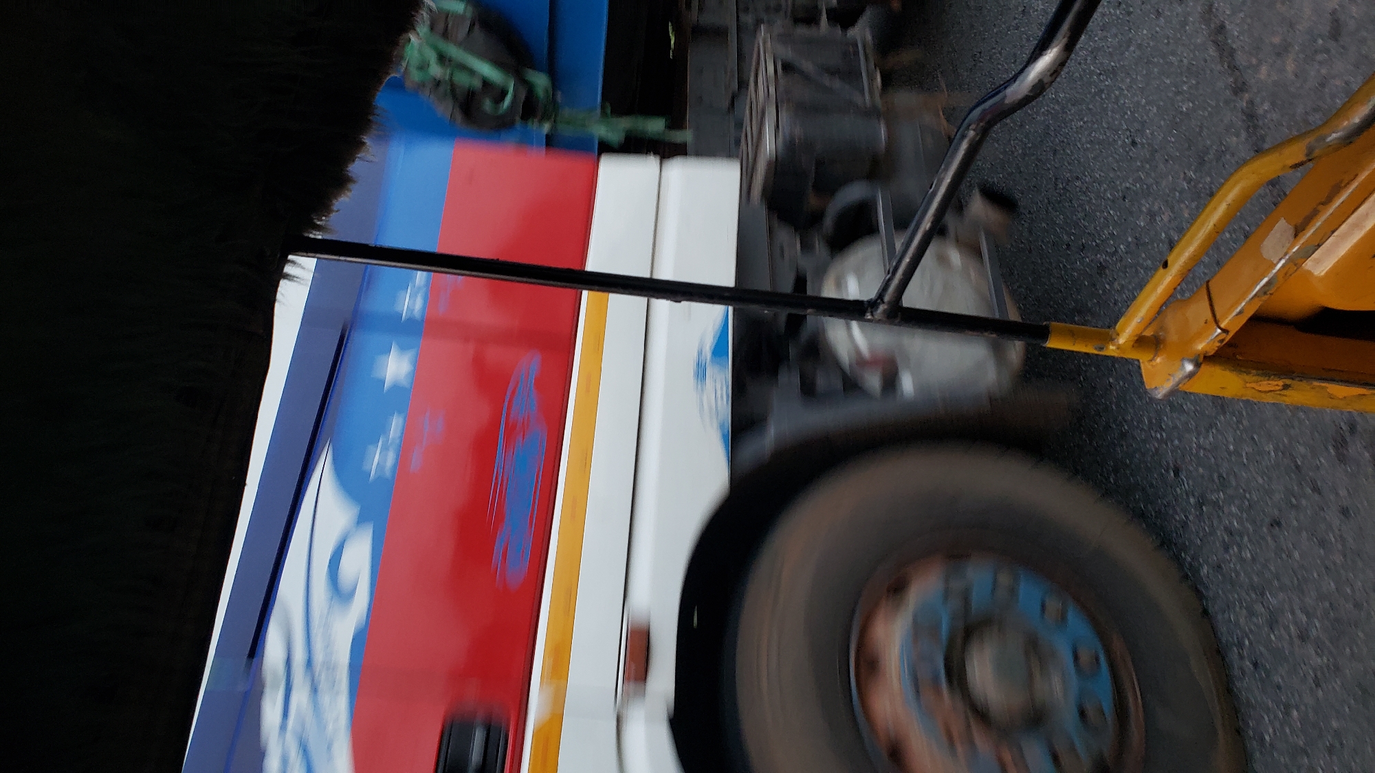 The image shows a side view of a moving truck with a red, white, and blue design on its side, including white stars. A large, blurred tire of the truck is visible on the left. In the foreground, a yellow object, possibly part of a vehicle or equipment, with black metal bars is also visible and blurred due to motion. The scene appears to be on an asphalt road, suggesting an urban or suburban setting. The overall impression is that the photo was taken from another moving vehicle. Given the prompt specified Jalingo, Nigeria, this scene likely depicts a street or highway within the city.