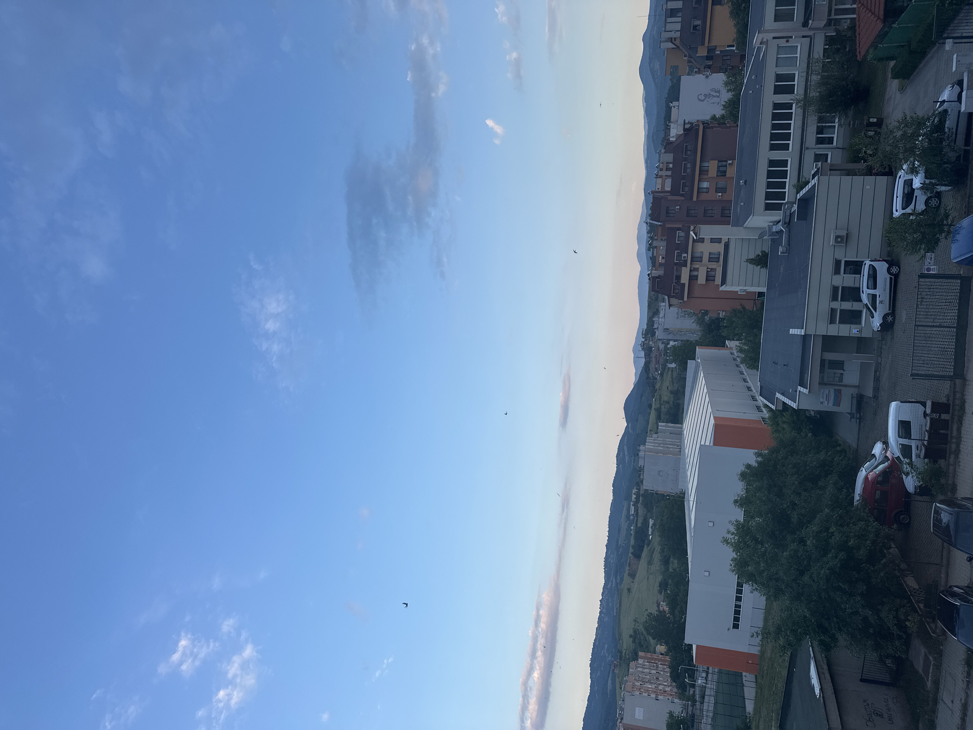 The image captures a panoramic view of an urban area in Blagoevgrad, Bulgaria, under a clear blue sky with scattered white and light gray stratocumulus clouds. The lighting suggests late afternoon or early evening. Multiple small, dark birds are visible flying across the sky.
In the mid-ground and background, a range of green, tree-covered hills and mountains defines the horizon, receding into a light atmospheric haze.
The urban landscape is dominated by a mix of residential and institutional buildings. On the left side, several multi-story apartment buildings with neutral facades (grey, white, beige) are visible. In the center-left, a large, modern sports hall or industrial building with a white main structure and distinctive orange-sloped sections is prominent, featuring a multi-paneled roof. Adjacent to it on the left is a green artificial turf field.
To the right of the sports hall, more residential buildings with varying architectural styles, including terracotta roofs and multi-colored facades (brown, beige), are present. One building on the far right has a large mural depicting a human figure on its side.
In the foreground, dense green trees partially obscure parts of the scene. To the right of these trees is a smaller, institutional-looking building complex with a grey facade and flat roofs, featuring numerous windows. A sign on the side of this building clearly displays the Cyrillic word "Общински" (Obshchinski, meaning Municipal) and the colors of the Bulgarian flag, indicating a municipal function.
A fenced parking area is located in front of the municipal building and extends to the left. Several vehicles are parked there, including a red minivan/van, two white vans (one appears to be a Dacia Dokker), and parts of two dark-colored cars/SUVs. Other vehicles are sparingly visible on streets or parking areas throughout the scene. No people are discernible in the image.