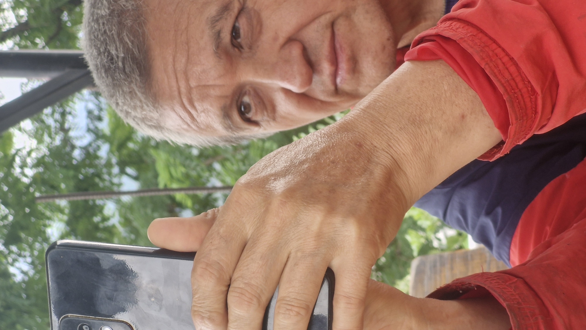 A close-up, eye-level shot shows a person holding a dark smartphone horizontally with their left hand, as if taking a selfie. The person's face is partially visible in the right side of the frame, with their gaze directed upwards and to the right. They have grey hair and visible wrinkles on their forehead and around their eyes. They are wearing a red jacket with navy blue sections on the torso and sleeves. The background is blurred, with green foliage and hints of what might be a light-colored sky or structure. The lighting suggests it is daytime. The image is an outdoor shot, possibly in a park or garden setting, with no specific landmarks of Vashisht, India, being identifiable from the visual evidence.
