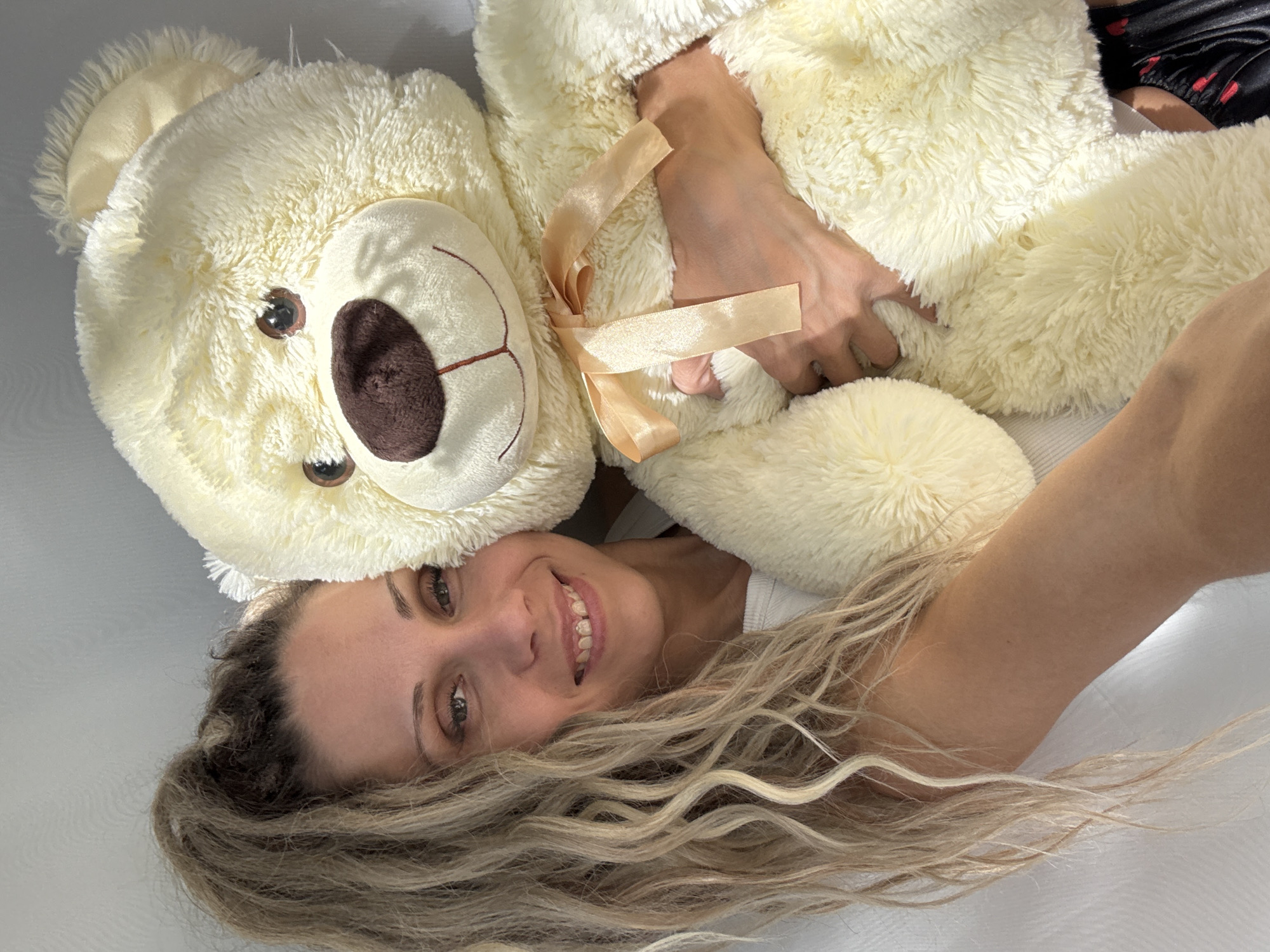 A woman with blonde, curly hair pulled into a high ponytail is holding a large, cream-colored teddy bear. She is smiling and looking at the camera, with her left arm wrapped around the bear. The teddy bear has a brown nose and embroidered smile, and is wearing a beige ribbon tied in a bow around its neck. The background is a plain, light-colored wall. The scene is indoors, with soft lighting. The image does not provide any information about the location in the city of Zhukovo, Russia.