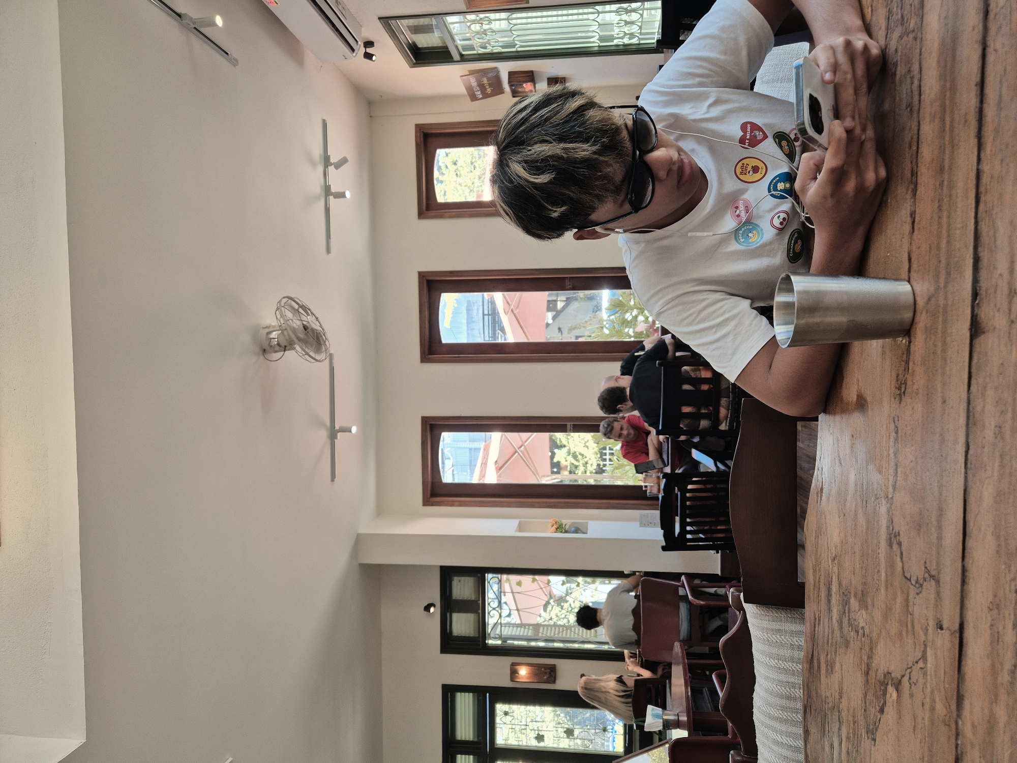 This is a medium shot of a young person sitting at a wooden table in a cafe in Da Nang, Vietnam. The person is wearing a white t-shirt with cartoon character patches and sunglasses. They are holding a smartphone and looking down at it, with earbuds in their ears. A metal cup is on the table to their left. In the background, other patrons are visible at tables, and large windows look out onto an outdoor area with trees and umbrellas. The cafe appears to be well-lit with natural light and ceiling fans and track lighting. The decor is modern with exposed brick and wooden furniture.