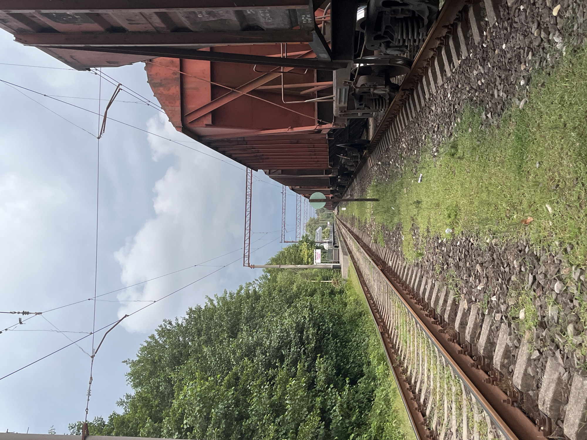 The image depicts a railway line in the city of Ureki, Georgia. On the right side of the frame, a long freight train composed of red hopper cars is parked on the tracks. The train cars are large and appear to be carrying loose material. The railway tracks themselves are visible, with concrete sleepers and ballast. To the left of the tracks, a dense line of green trees forms a backdrop, suggesting a natural or semi-rural environment. In the distance, a small railway station platform can be seen, with a sign that reads "UR EK I" (Ureki). Overhead, power lines for the electric train system stretch across the sky, which is overcast with clouds. The overall scene conveys a sense of a functional, working railway infrastructure within a relatively green and open area. There are no people visible in the image.