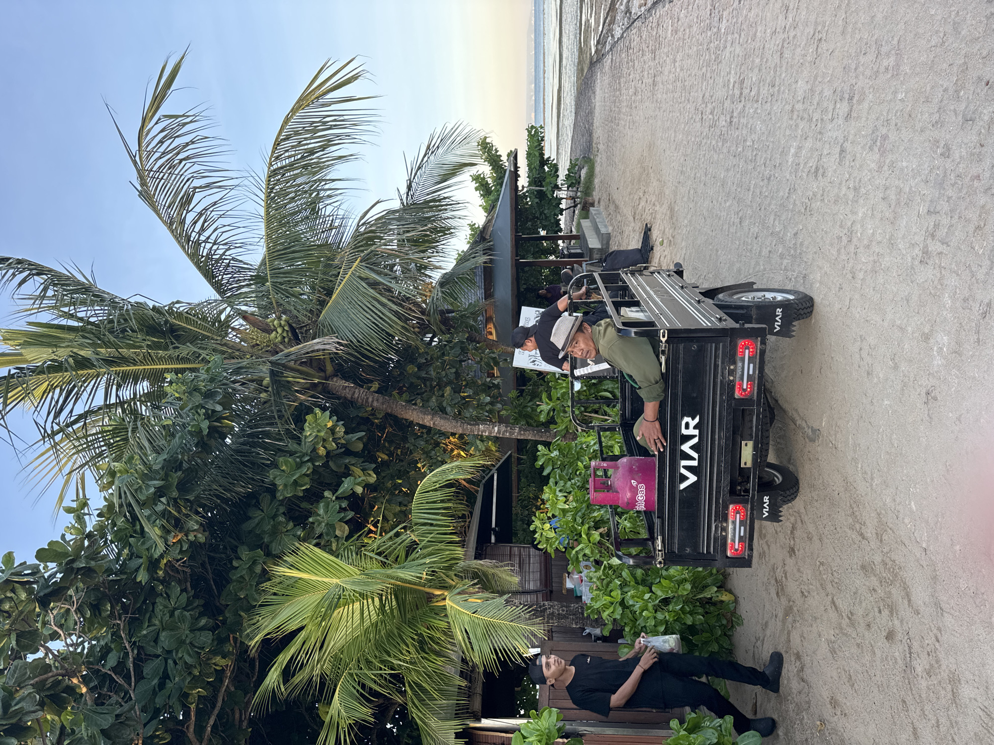 The image shows a scene at the beach. A palm tree is in the left of the image. There are two men and one is sat on a motorized tricycle. In the back of the tricycle is a pink gas tank. The other man is dressed in all black. In the background, the sea is visible.
