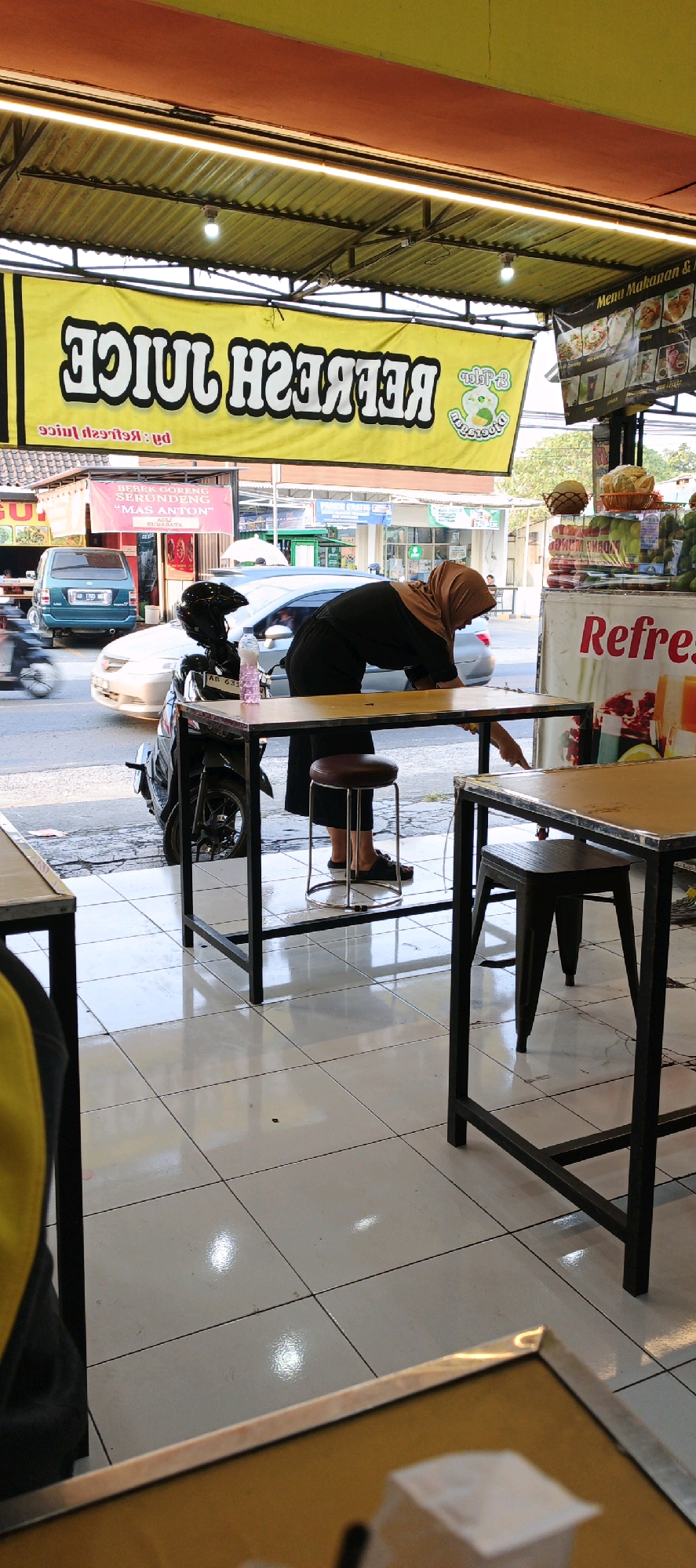 In the city of Sidoarum, Indonesia, a person wearing a hijab and a black outfit is bending down at a table outside a juice stand. The sign above the stand reads "REFRESH JUICE" in large, bold letters. The stand appears to be on a street, as a motorcycle and cars are visible in the background. The flooring is tiled and reflective, indicating an indoor/outdoor setting. The scene is brightly lit, likely by natural sunlight and overhead lights.