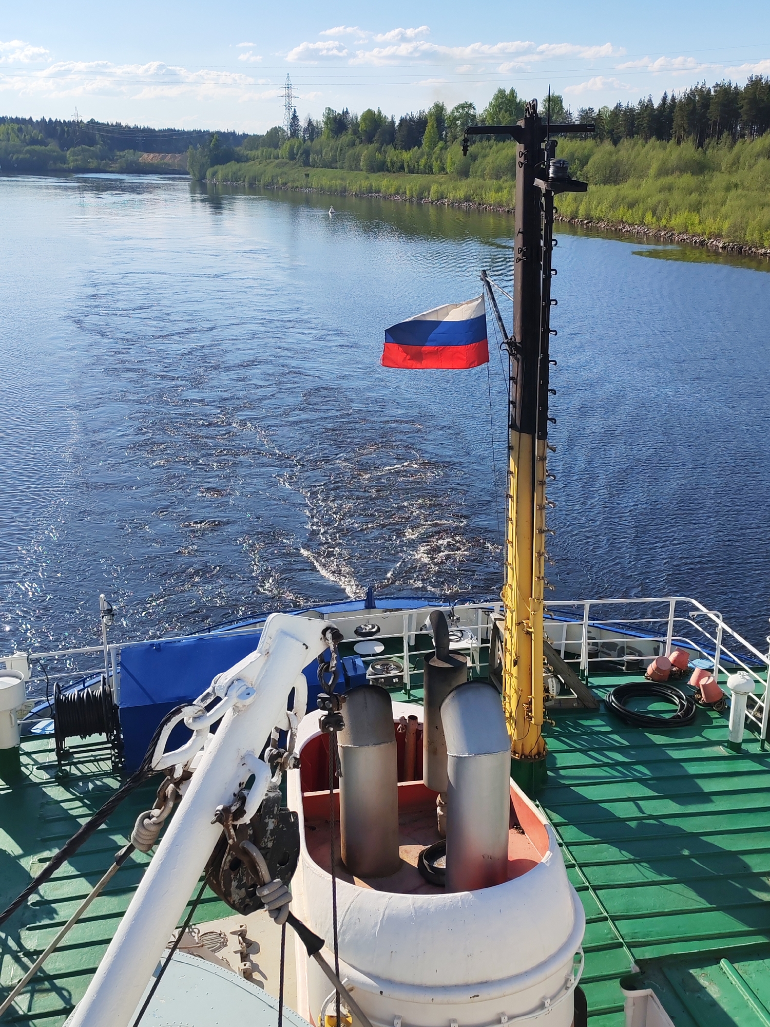 The image shows the deck of a riverboat, possibly a cruise ship, moving through a waterway in Depo, Russia. The Russian flag is prominently displayed. The stern of the boat creates a foamy wake in the blue water. The shore is lined with green vegetation and power lines are visible in the background. The deck of the boat is green with a white railing. A white and red structure with cylindrical exhaust pipes is visible in the foreground, along with white mechanical equipment.
