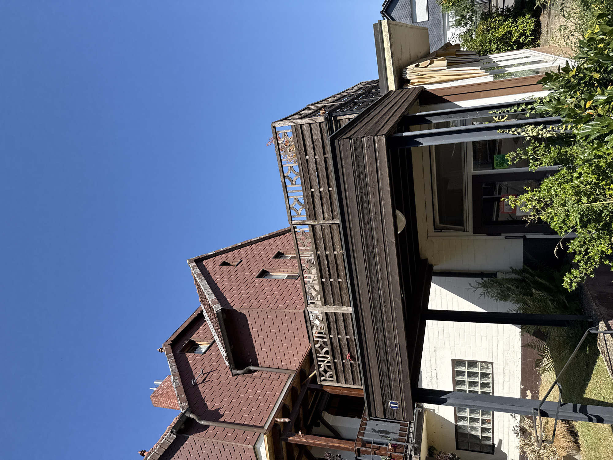 The image depicts a two-story building with a distinct architectural style, set against a clear blue sky. The upper story is clad in reddish-brown tiles, with a pitched roof and a prominent brick chimney. A wooden balcony with a decorative railing spans part of this level. Below, the ground floor features a white brick facade with a dark brown, sloped roof structure supported by dark metal beams. This lower section has large windows, some made of glass blocks, and an awning partially covering the area. Lush green foliage and grasses are visible in the foreground, suggesting an outdoor setting. The building appears to be located in a residential or commercial area, as indicated by the presence of other buildings in the background. The number "1" is visible on the lower section of the building, possibly indicating a unit or entrance number. The overall scene is well-lit by sunlight, casting shadows on the building and surrounding vegetation. The location is identified as Bad Sachsa, Germany, a town known for its spa and recreational facilities, often characterized by a mix of traditional and modern architecture.