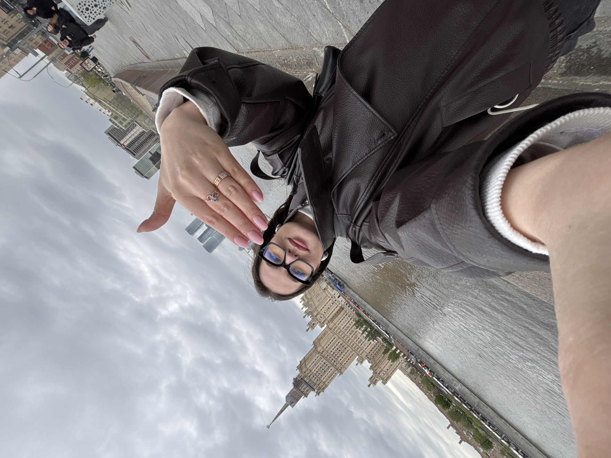 The image is a selfie taken by a person wearing glasses, a white sweater, and a brown leather jacket. The person is standing on a sidewalk made of grey pavers and is holding their hand up, showing off two rings. In the background, there is a body of water and a large Stalinist-era skyscraper in Moscow, Russia. The sky is cloudy. Other people can be seen in the background on the sidewalk.