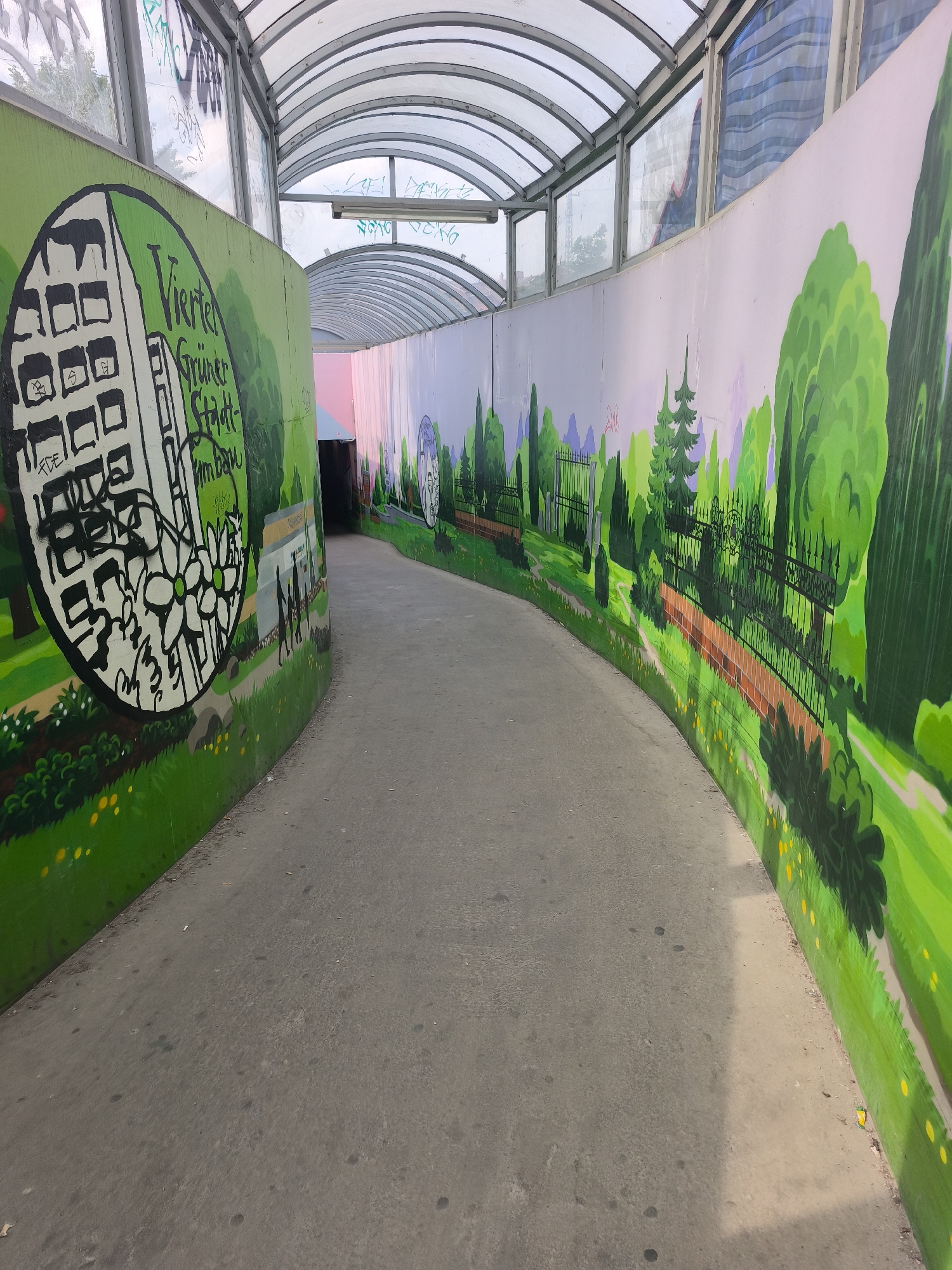 The image captures an interior shot of an underpass or tunnel in Vienna, Austria. The tunnel has a curved ceiling constructed with transparent or translucent panels, likely made of plastic or glass. The walls of the tunnel are adorned with vibrant murals, primarily featuring green landscapes, trees, and urban elements. On the left wall, there is a large circular mural with a building and a floral design. The murals on the right wall depict a park with trees, benches, and a wrought iron fence. The floor of the tunnel is made of concrete and appears to be worn. The tunnel's opening suggests it is a pedestrian or bicycle passage.