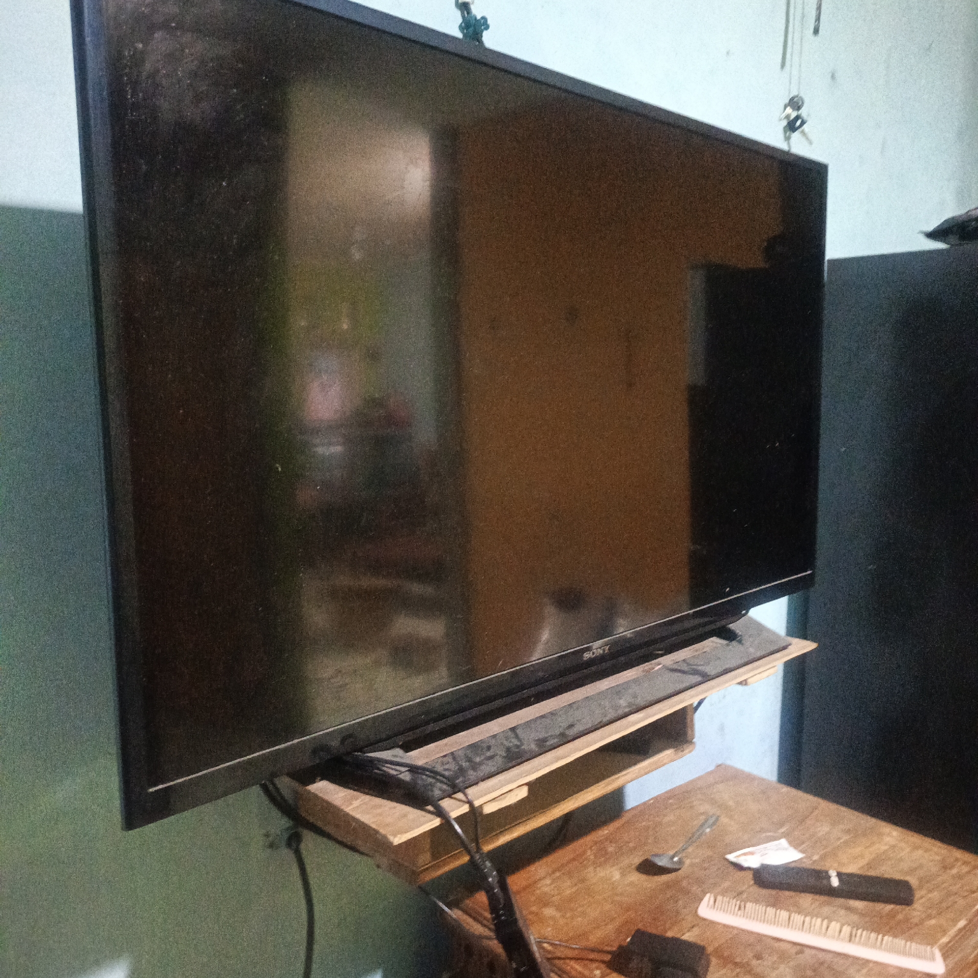 A Sony television is mounted on a wall and is resting on a wooden shelf. A spoon, a small white packet, a comb, and a dark object are on a wooden table below the shelf. There is a black cord connected to the television. The wall behind the television is painted a light blue. A dark speaker is to the right of the television. The scene appears to be in a room in Karangtuang, Indonesia.