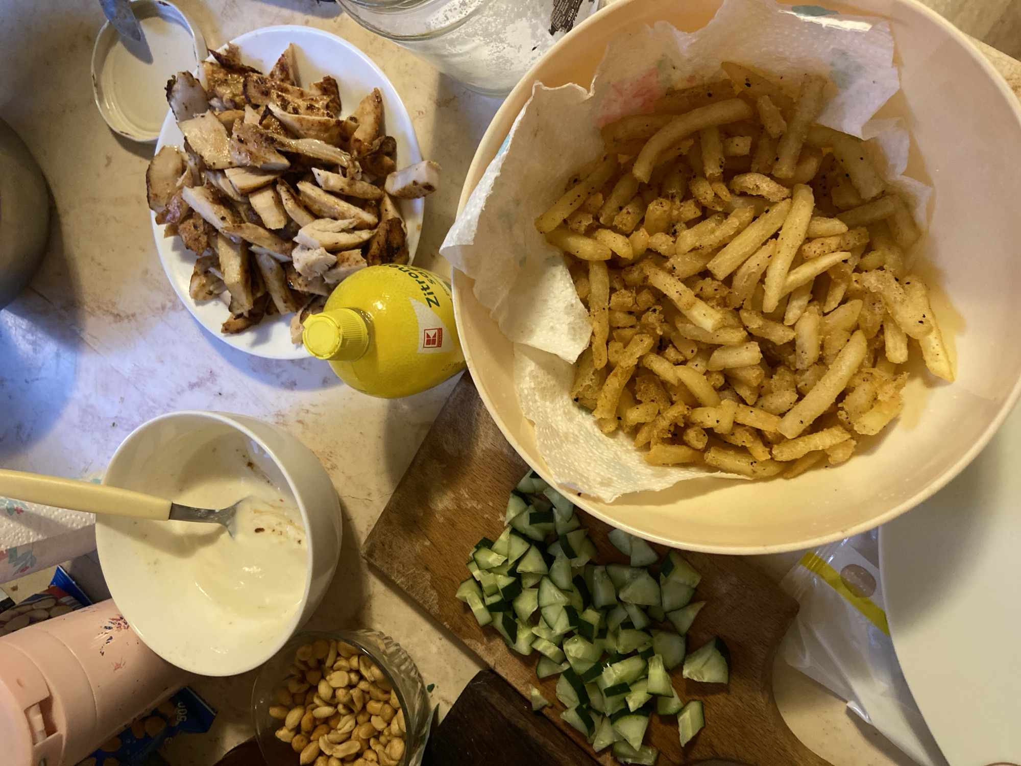 A close-up, overhead shot shows food preparation on a light-colored countertop, possibly in a home kitchen in Mindia, Bulgaria. In the foreground, a bowl contains a generous portion of golden french fries, drained on a paper towel lining the beige bowl. To the left, a wooden cutting board displays a pile of diced cucumbers. Above the cucumbers, a glass bowl holds a serving of salted peanuts. Further back, a white bowl contains a creamy white sauce with a spoon resting inside. To the right of the sauce bowl, a white plate is mounded with sliced, grilled chicken. A bright yellow bottle with a red label and a silver cap sits in front of the chicken, and a glass jar filled with white granular material, likely salt, is visible to the right of the plate. Other objects, including a pink thermos and a paper towel dispenser, are partially visible in the background. The overall scene depicts ingredients and a prepared dish, suggesting a meal is about to be assembled or served.