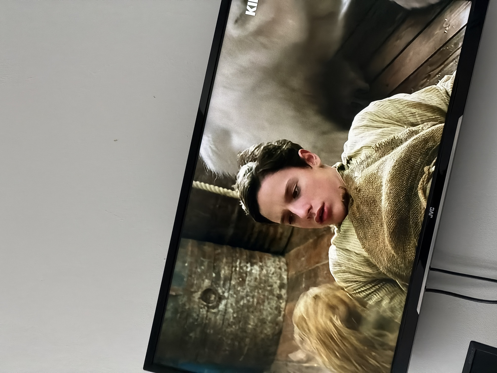 The image shows a JVC television displaying a scene with a young man wearing a coarse, possibly woolen, garment. The background suggests a rustic setting, possibly a barn or a similar wooden structure. Ropes are also visible. The location is not discernible from the image; therefore, determining its location in Sofia, Bulgaria, is impossible.