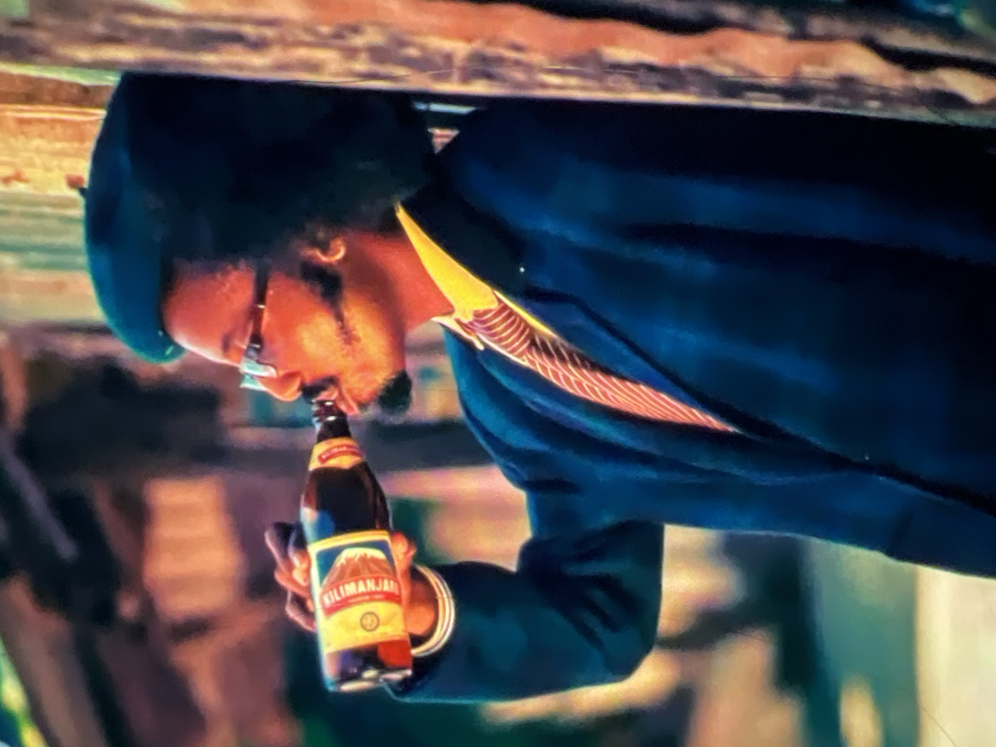 A man wearing a dark beret, glasses, a yellow collared shirt, a striped tie, and a dark blue jacket is drinking from a brown glass bottle. He is leaning against a wooden post with his arm raised to his mouth. The background is blurred, with hints of orange and green structures, suggesting an outdoor setting, possibly a market or street scene in Lagos, Nigeria. The man has dark, curly hair and a goatee. He is also wearing a hoop earring in his left ear. The label on the bottle is partially visible and appears to be for Kilimanjaro.