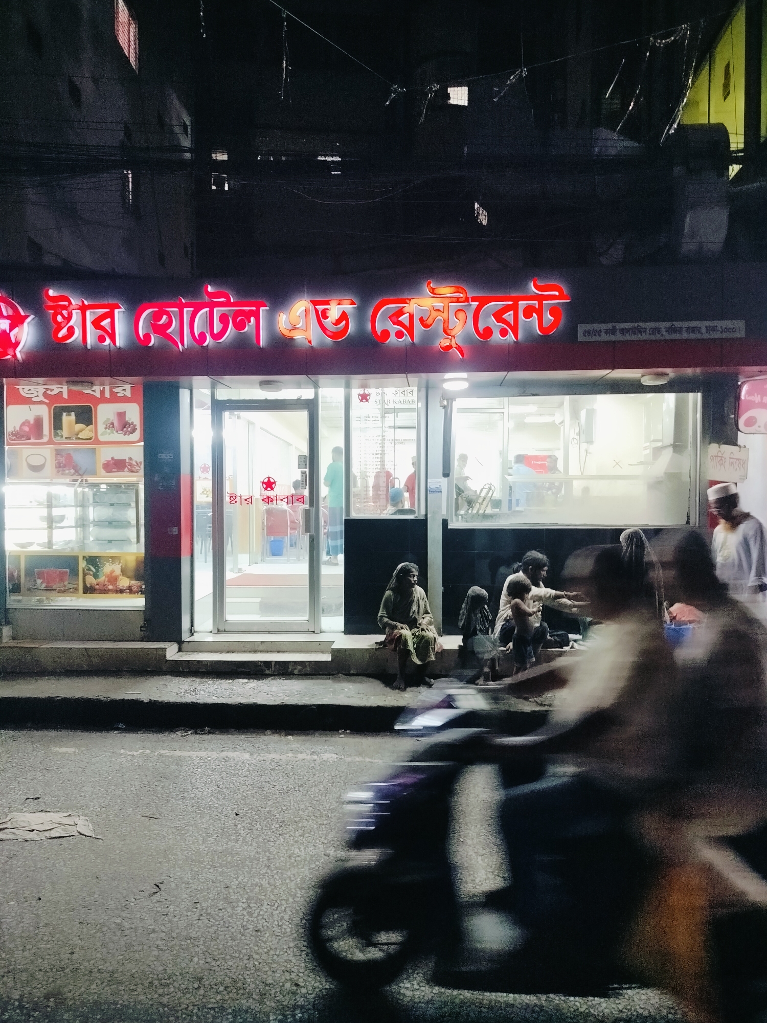 The image captures a street-level view of a brightly lit restaurant and hotel facade at night in Dhaka, Bangladesh. A prominent red neon sign reads "স্টার হোটেল এন্ড রেস্টুরেন্ট" (Star Hotel and Restaurant). Below the main sign, another smaller red sign states "স্টার কাবাব" (Star Kabab). The entrance to the establishment features large glass doors, revealing a well-lit interior with tables and chairs.
In front of the restaurant, several people are seated on the ground. A blurred image of a motorcycle in motion suggests activity on the street. The street surface is visible, with some scattered debris. The overall scene is a nighttime urban environment.
