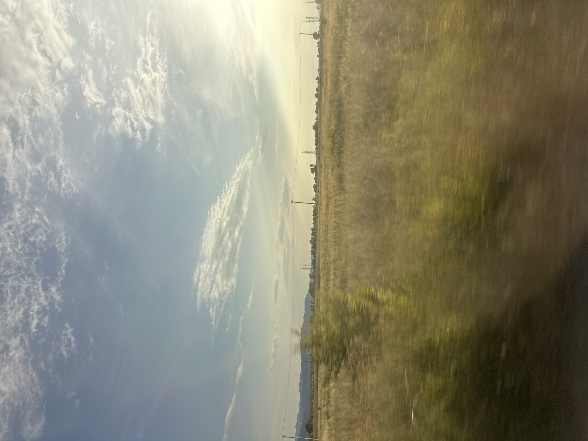 This is a photograph of a vast, open landscape taken from a moving vehicle, likely a train or car, given the motion blur. The image is oriented vertically, with the sky occupying the left two-thirds and the ground the right third.
The sky is a pale blue, filled with wispy, white clouds that are illuminated by the sun, suggesting it is either early morning or late afternoon. The sun itself is not directly visible but its bright light is casting a soft glow across the sky, creating a slightly hazy and serene atmosphere.
The ground consists of a dry, grassy field that stretches to the horizon. The grass is a muted yellow and green, reflecting the dry conditions. Scattered throughout the field are occasional trees, appearing as dark vertical smudges due to the motion blur. In the distance, a range of low hills can be seen, fading into a light haze. Power lines and their tall, lattice towers are visible in the middle ground, stretching across the field and indicating the presence of human infrastructure.
The overall impression is one of a quiet, rural journey through a dry, expansive landscape under a bright, cloudy sky. The motion blur contributes to a sense of movement and passage. There is no visible text in the image. The location context of Sayanogorsk, Russia, suggests a potentially remote or steppe-like environment, consistent with the visual elements.