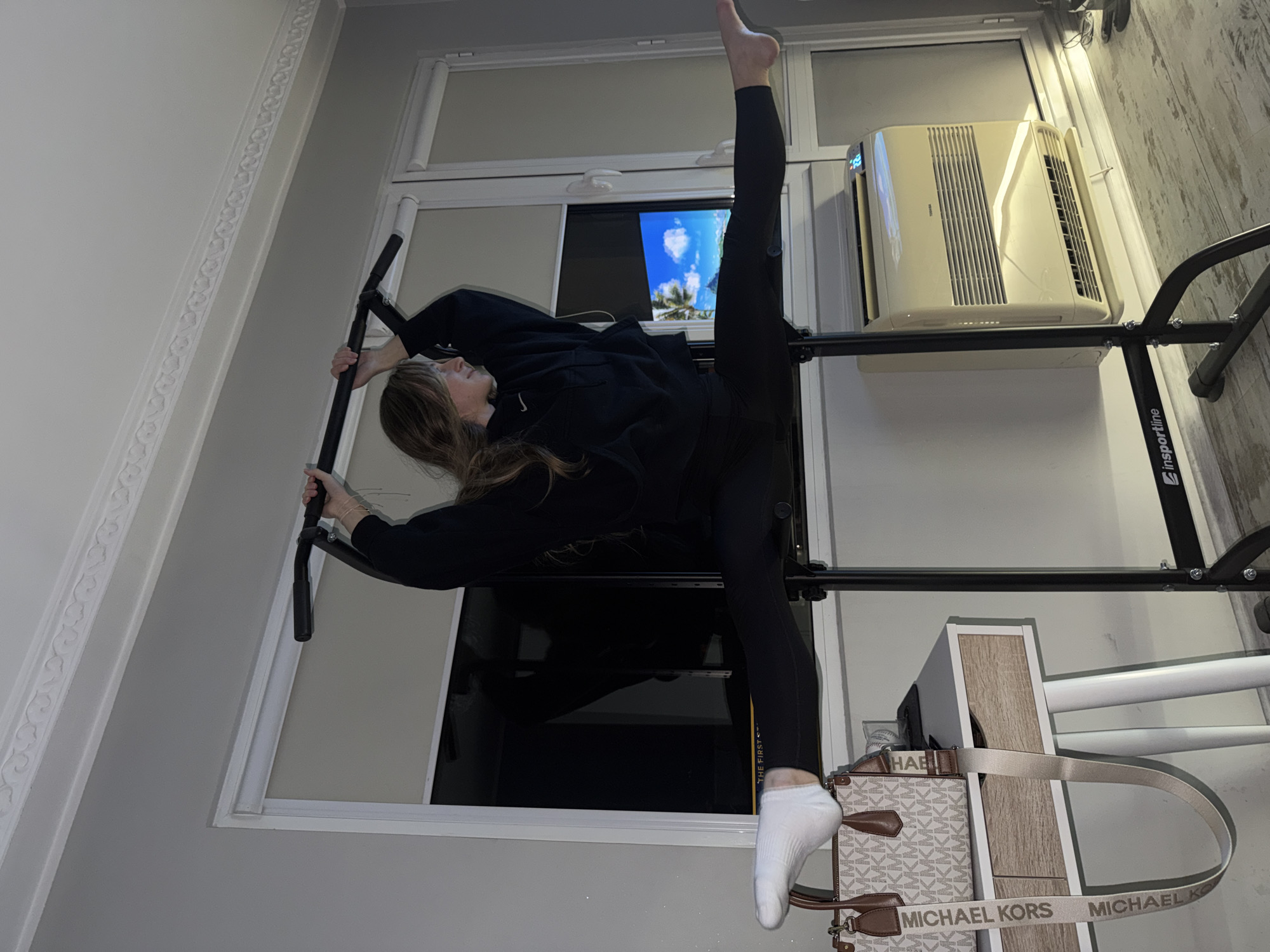A young woman with long brown hair is performing a split while holding onto a pull-up bar. She is wearing a black sweatshirt and black leggings, and one white sock on her raised foot. The pull-up bar is positioned in a window frame, with an air conditioning unit installed above it. A Michael Kors purse is hanging nearby. A television screen is visible in the window behind the woman. The room has light gray walls with decorative molding near the ceiling. The location is likely not in Plovdiv, Bulgaria, as there is no evidence to support this claim within the image.