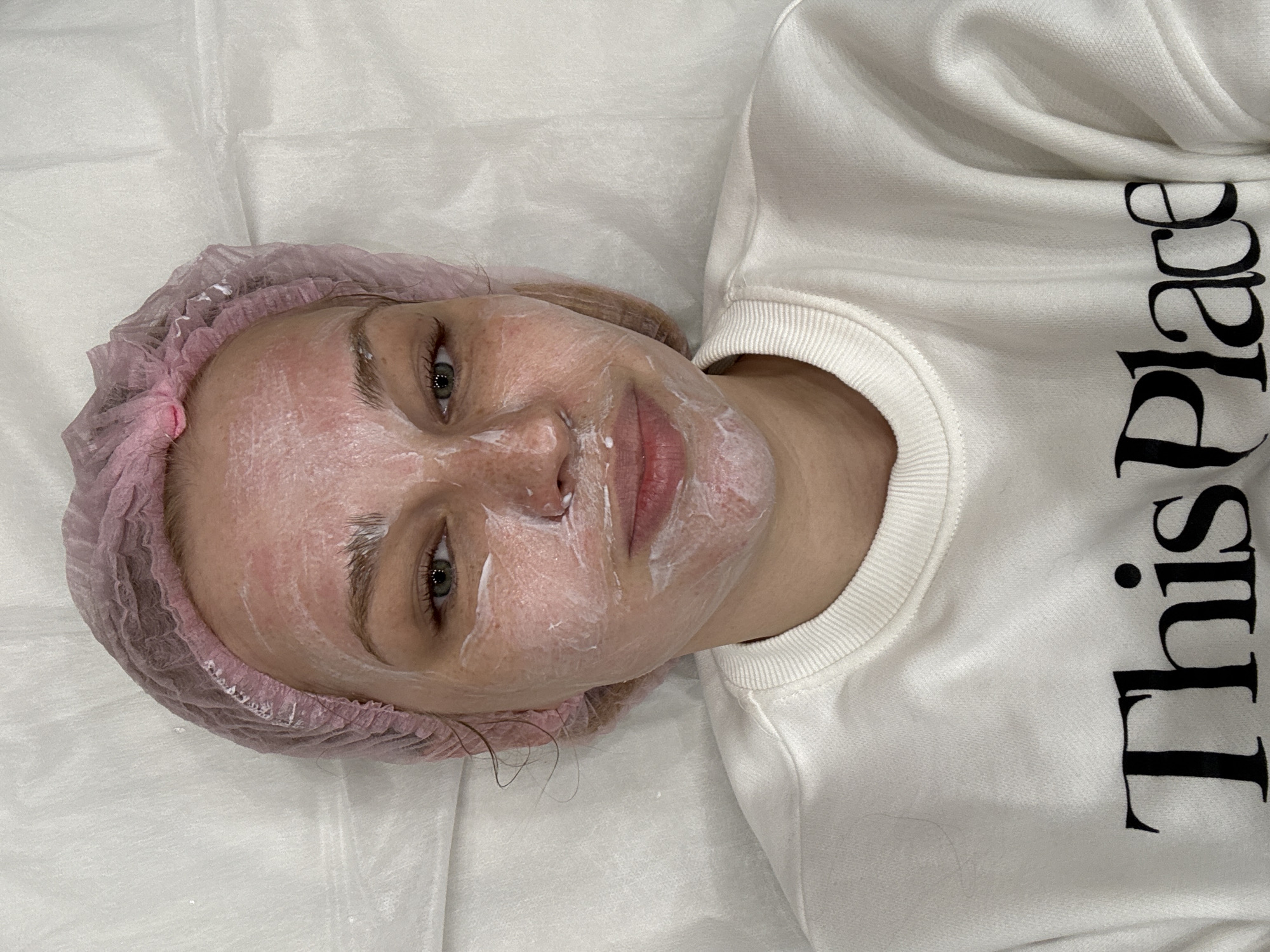 A young woman lies on a treatment bed covered with a white sheet, wearing a pink disposable hair net and a white sweatshirt with the phrase "This Place" printed vertically in black letters. Her face is covered in a white cream, indicative of a facial treatment. The context suggests a beauty salon or spa, but the specific location in Rusanovo, Russia is not discernible from the image alone.