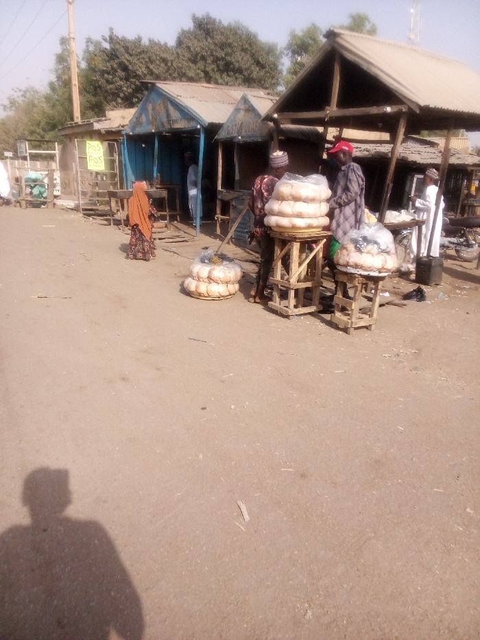 The image depicts an outdoor market or street scene in Dakata, Nigeria, bathed in bright, clear daylight, as evidenced by the distinct shadow of the photographer in the bottom left foreground. The ground is unpaved and appears dusty.
The central focus of the scene is the sale of numerous large, round loaves of bread, likely a local staple. These loaves are neatly stacked on rustic wooden stands of varying heights, and some are also placed on the ground, many covered in clear plastic bags for protection.
Several people are present in the mid-ground. On the left, a person wearing an orange or brown head covering and a patterned dress is seen from behind. Closer to the right, two men in traditional attire, one wearing a red cap, stand near the bread displays, seemingly involved in sales or purchases. Further back on the right, another person in white clothing is visible near a stall that appears to have a motorcycle or bicycle beside it. A person is also partially visible inside one of the background stalls.
In the background, a row of simple market stalls or shops with corrugated metal roofs are visible, some painted blue. A utility pole stands on the far left. A notable detail is a yellow sign hanging from a wooden stand on the left, which clearly displays the text "POS" repeated several times, indicating the availability of Point of Sale services. The scene conveys a typical, active market environment.