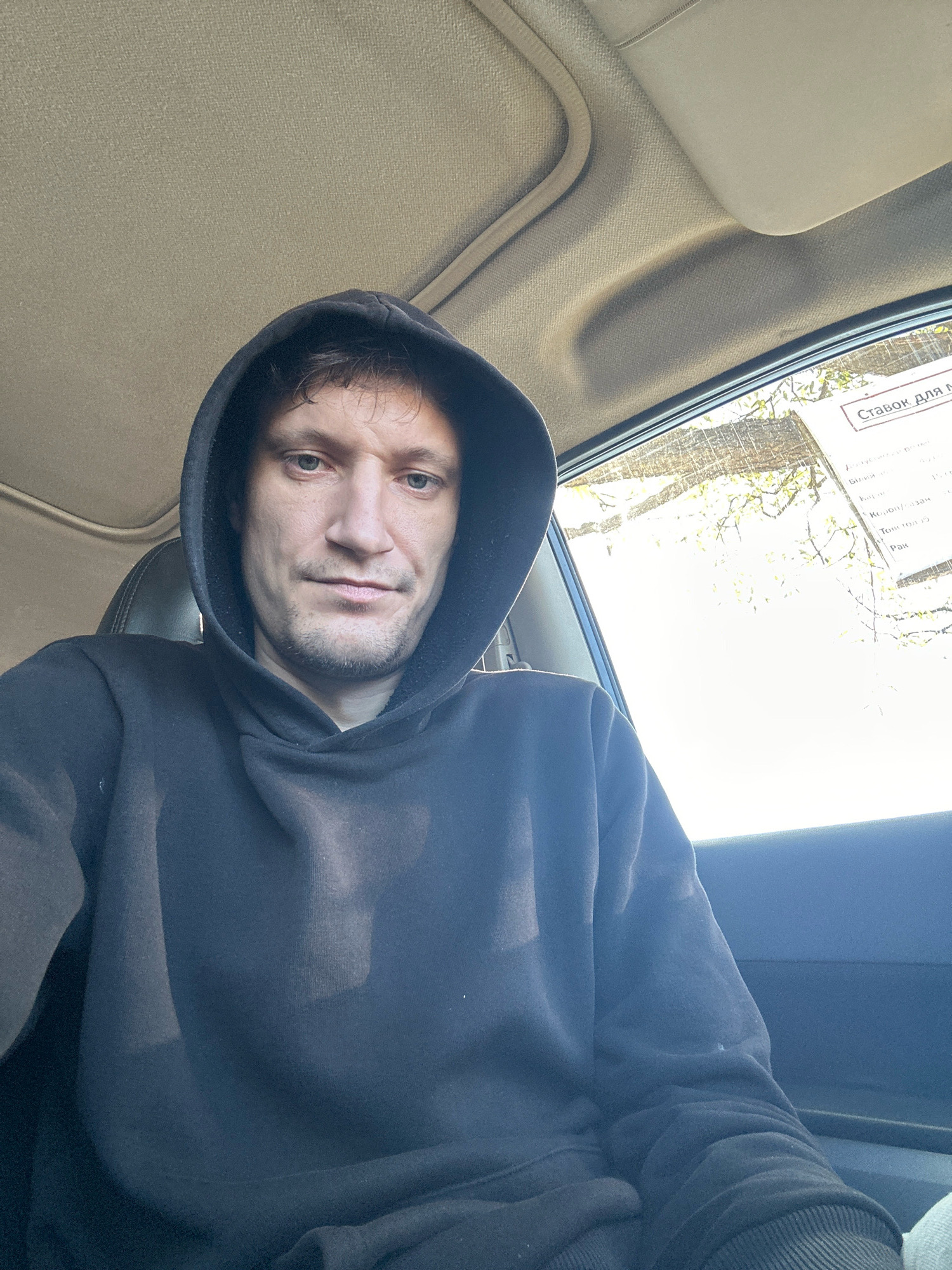 A man wearing a dark hoodie is sitting inside a car, looking towards the camera. He has light brown hair and stubble. The interior of the car is visible, with the ceiling and a portion of the window on the right. Outside the window, trees are visible against a bright sky. A document with text in Russian, possibly a receipt or a label, is affixed to the window. The image appears to have been taken during the daytime. There is no information in the image to suggest the location is Makiv, Ukraine.