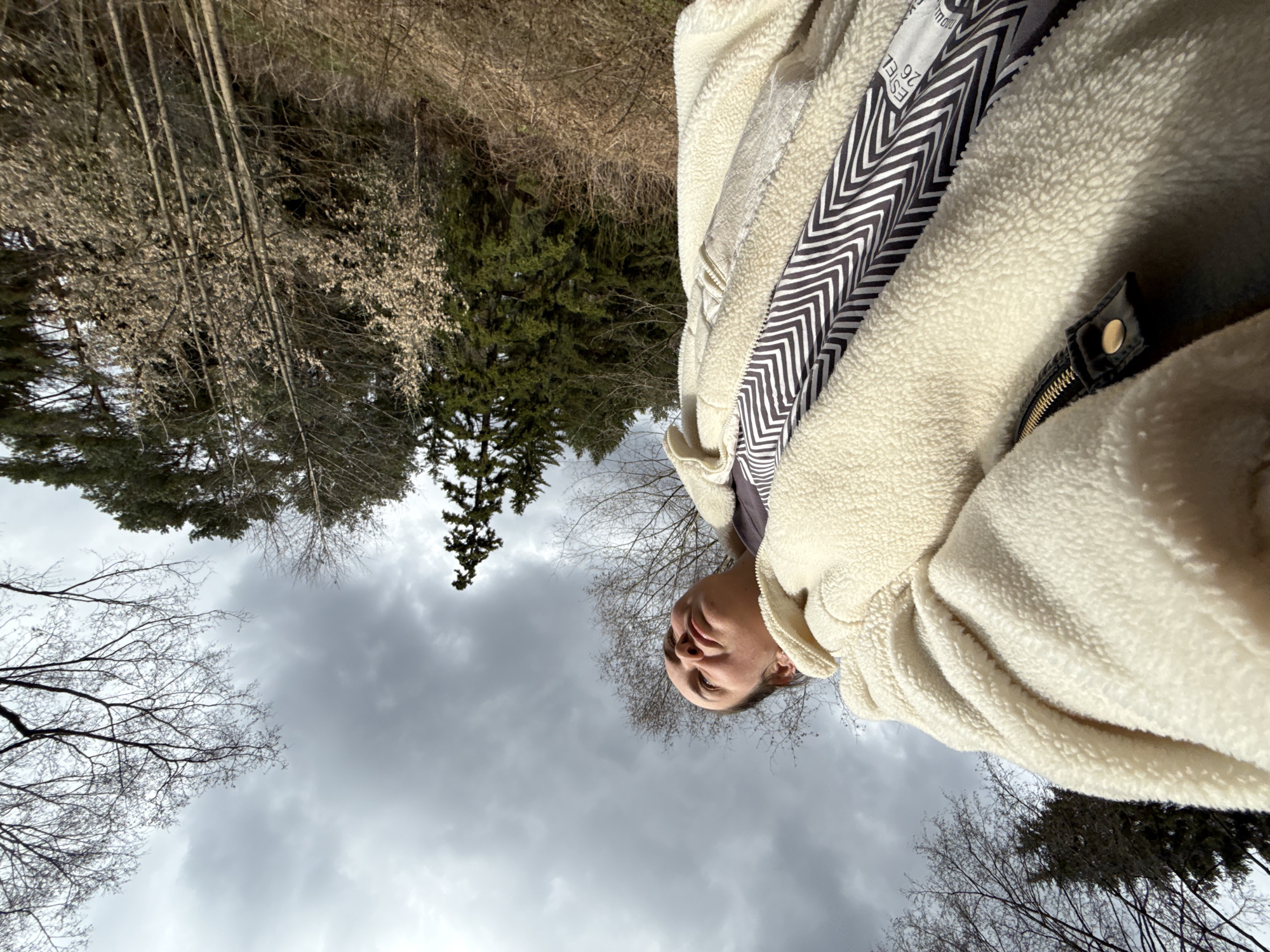 The image is a low-angle, upward-looking perspective shot of an individual in an outdoor, wooded setting, rotated 90 degrees clockwise. An adult, possibly female, with light skin and dark hair is visible in the top right quadrant, looking upwards with a neutral expression.
She is wearing a cream-colored, fluffy sherpa or fleece jacket over a black and white striped garment with a zigzag pattern. The jacket features a gold-toned zipper and a black strap with a brass snap button closure. A white clothing label is partially visible on the striped garment, displaying the text "ESTEL 26".
The background features a mix of bare deciduous trees, suggesting late winter or early spring, alongside dark green evergreen trees, indicating a forested or natural area. The sky is predominantly grey and overcast, contributing to a diffuse lighting condition. The scene depicts a moment of an individual in a natural, possibly tranquil, outdoor environment. The location context is Chaikovsky, Russia.