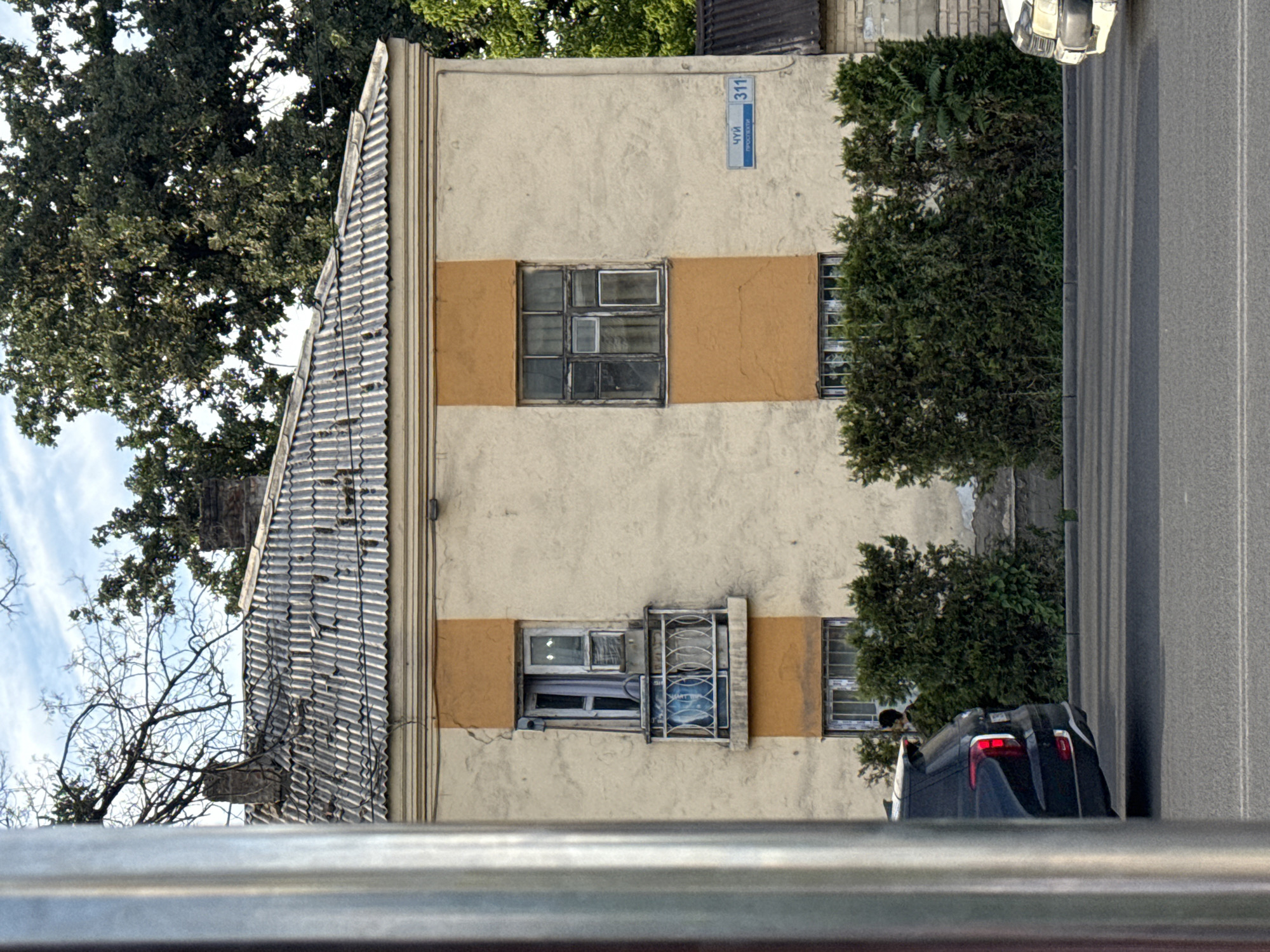 The image displays a portion of a light-colored building with a contrasting orange band running horizontally across its facade. The building has several windows. One prominent window on the orange band appears to be divided into a grid of smaller panes, some of which are dark or reflective. Below this, another window opening contains multiple panes and is partially covered by decorative metal bars. To the right of this, a smaller window is visible. The roof of the building is covered in corrugated metal tiles.
Above the orange band, a blue street sign is affixed to the building. The sign contains white text which appears to be Cyrillic. The number "311" is clearly visible at the top of the sign, and below it, text includes "ЧУЙ" and "ПРОСПЕКТ".
To the left of the building, large trees with dark green foliage are visible, as well as the bare branches of a deciduous tree against a bright sky with some clouds. To the right, a dark-colored vehicle is parked next to a paved road, which has visible lane markings. The orientation of the image is sideways. The scene suggests an outdoor setting, possibly a residential or commercial area, during daylight hours. There are no people clearly visible in the image.