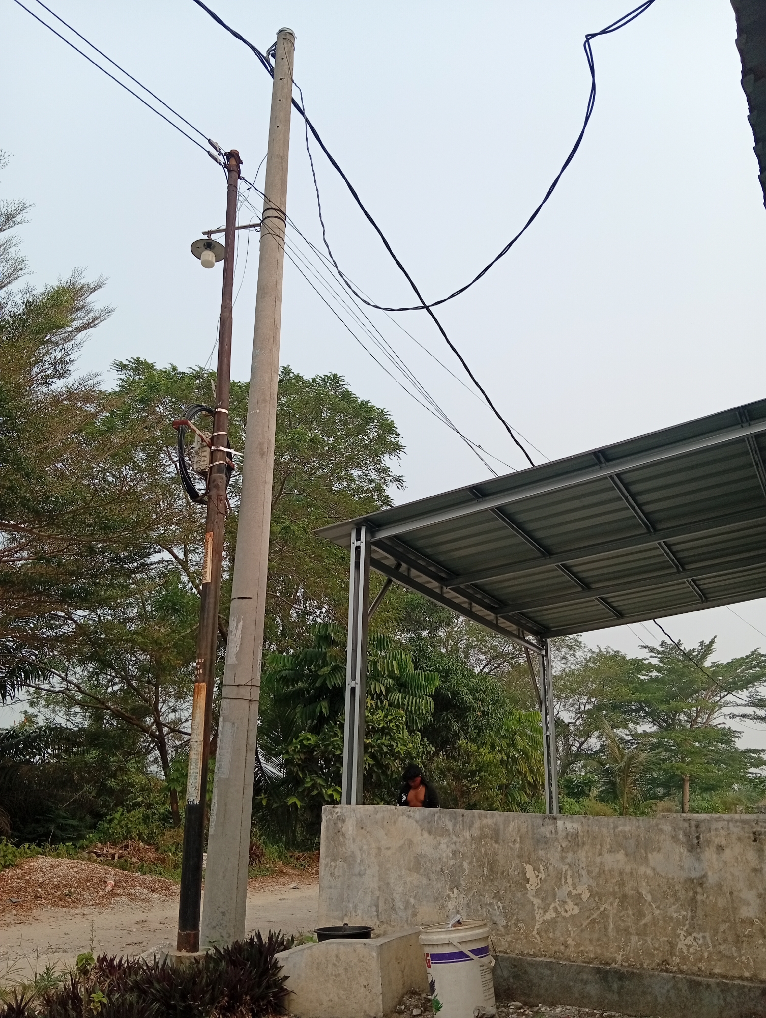 The image depicts an outdoor scene in Kualu, Indonesia, under an overcast sky. Dominating the foreground are two utility poles, one concrete and one rusty metal, with a street lamp attached to the metal pole. Electrical wires are strung between the poles and extend across the frame. To the right, a modern, grey metal-roofed structure is visible, supported by a concrete wall. A person, partially obscured, is seen near the structure, leaning over the concrete wall, with their torso visible. The background is filled with dense green foliage and trees, suggesting a semi-rural or natural setting. The ground appears to be a dirt path with scattered debris and some low-lying plants.